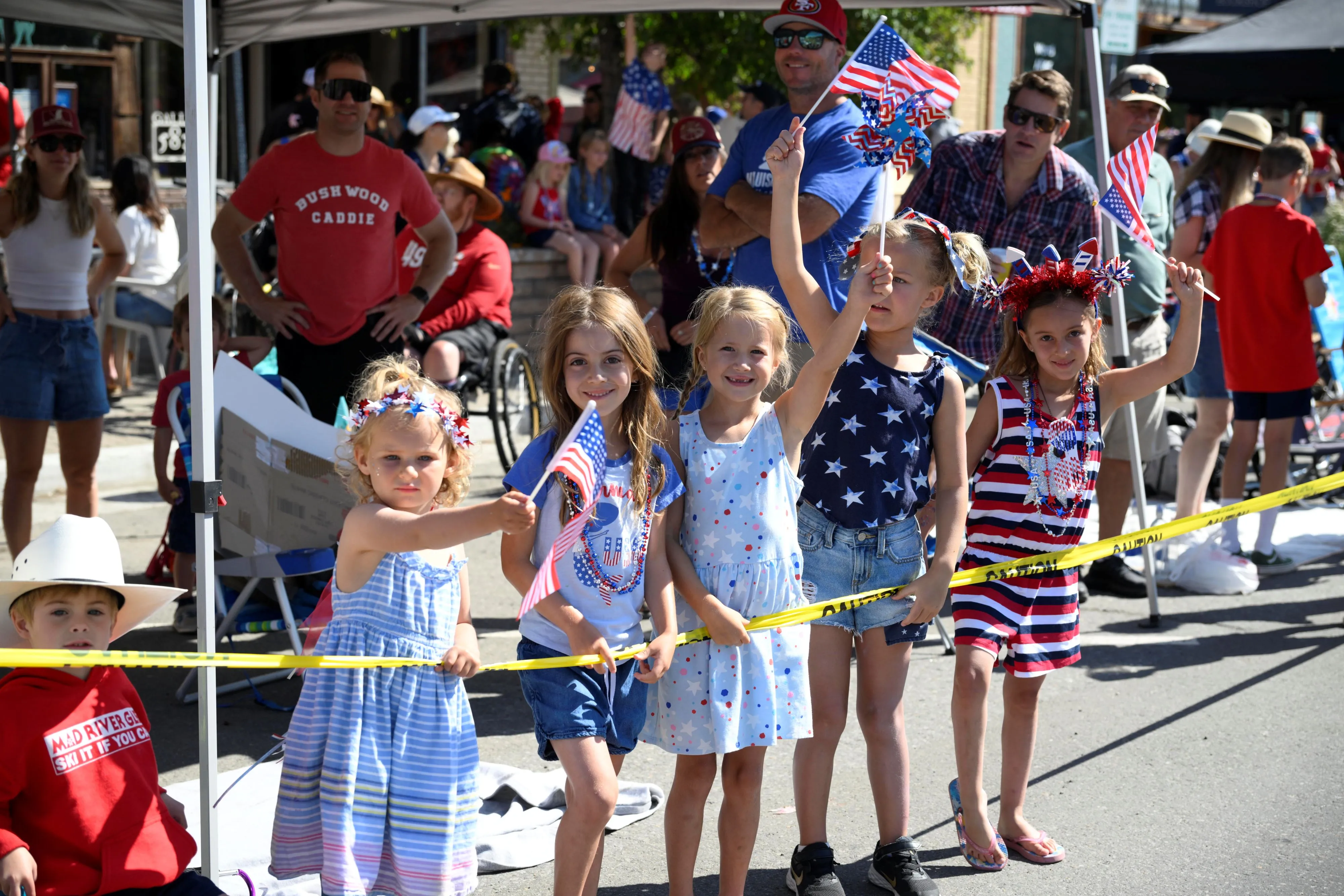 Group of five children holding yellow caution tape and American flags at an outdoor event, with adults and other people in the background.