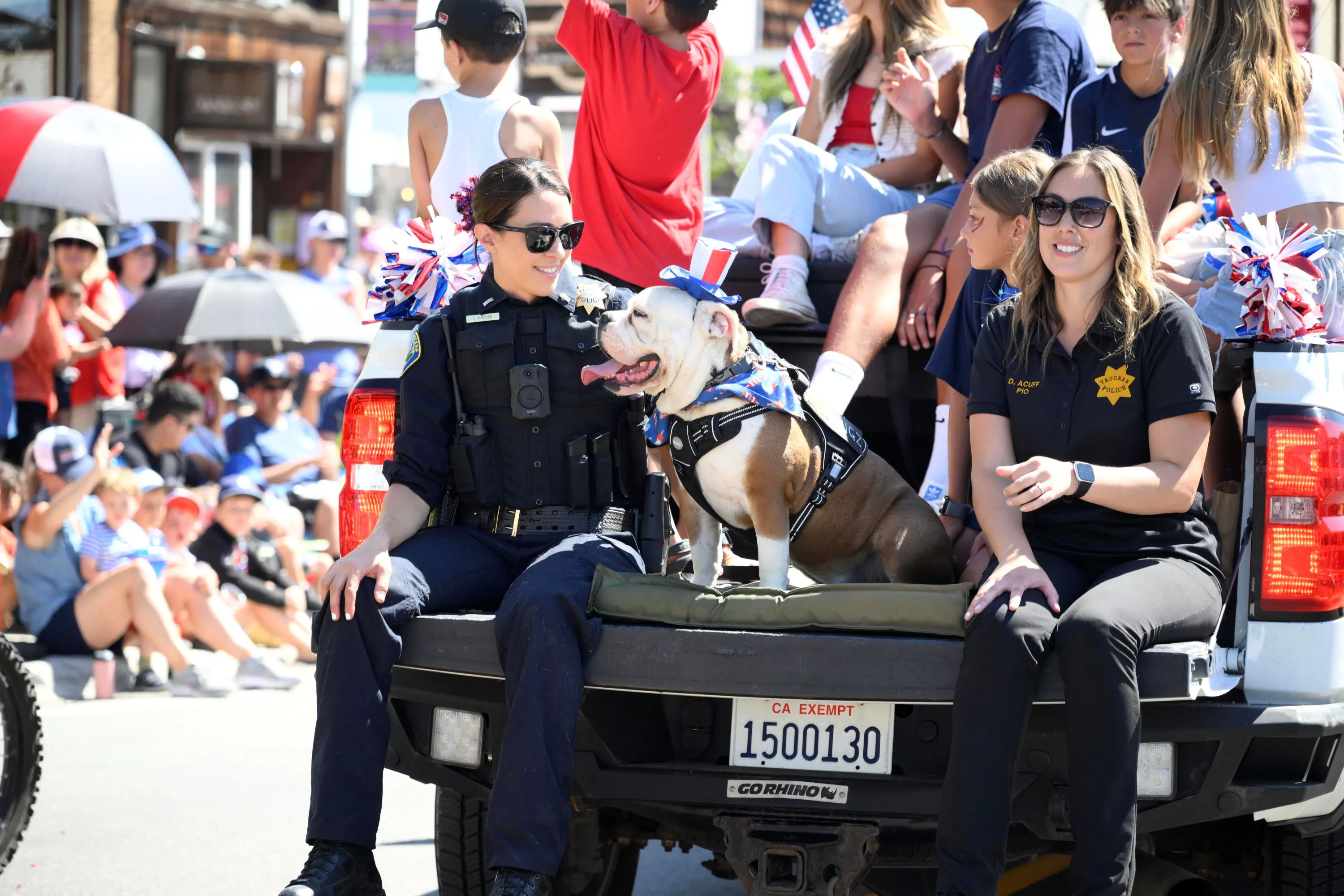 Two female police officers and a bulldog wearing a patriotic hat sitting on the back of a pickup truck during a street parade.