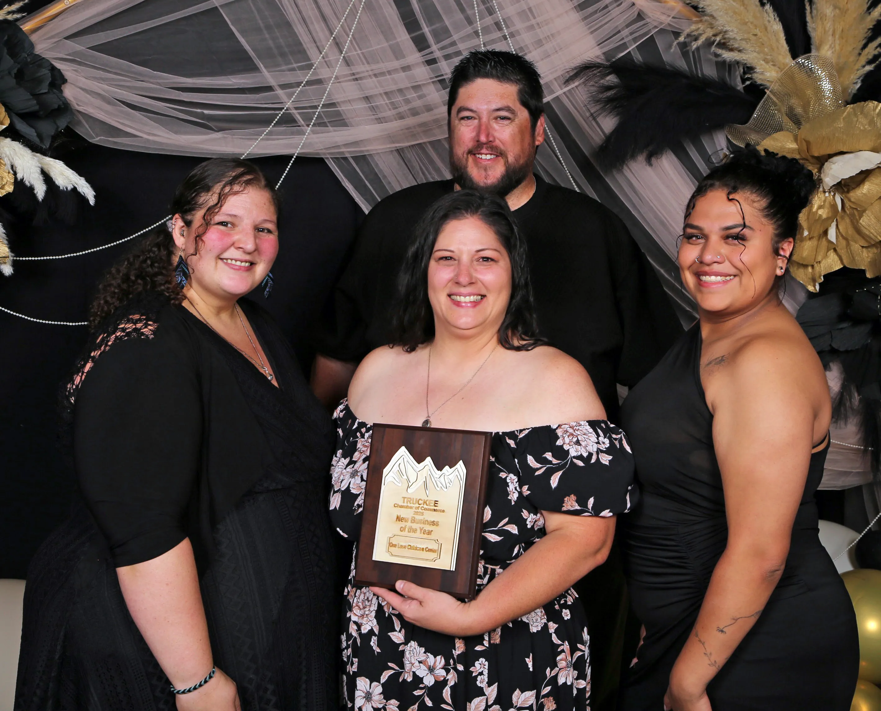 Four smiling people posing in front of a decorated backdrop, one holding a plaque that says 'New Business of the Year'.