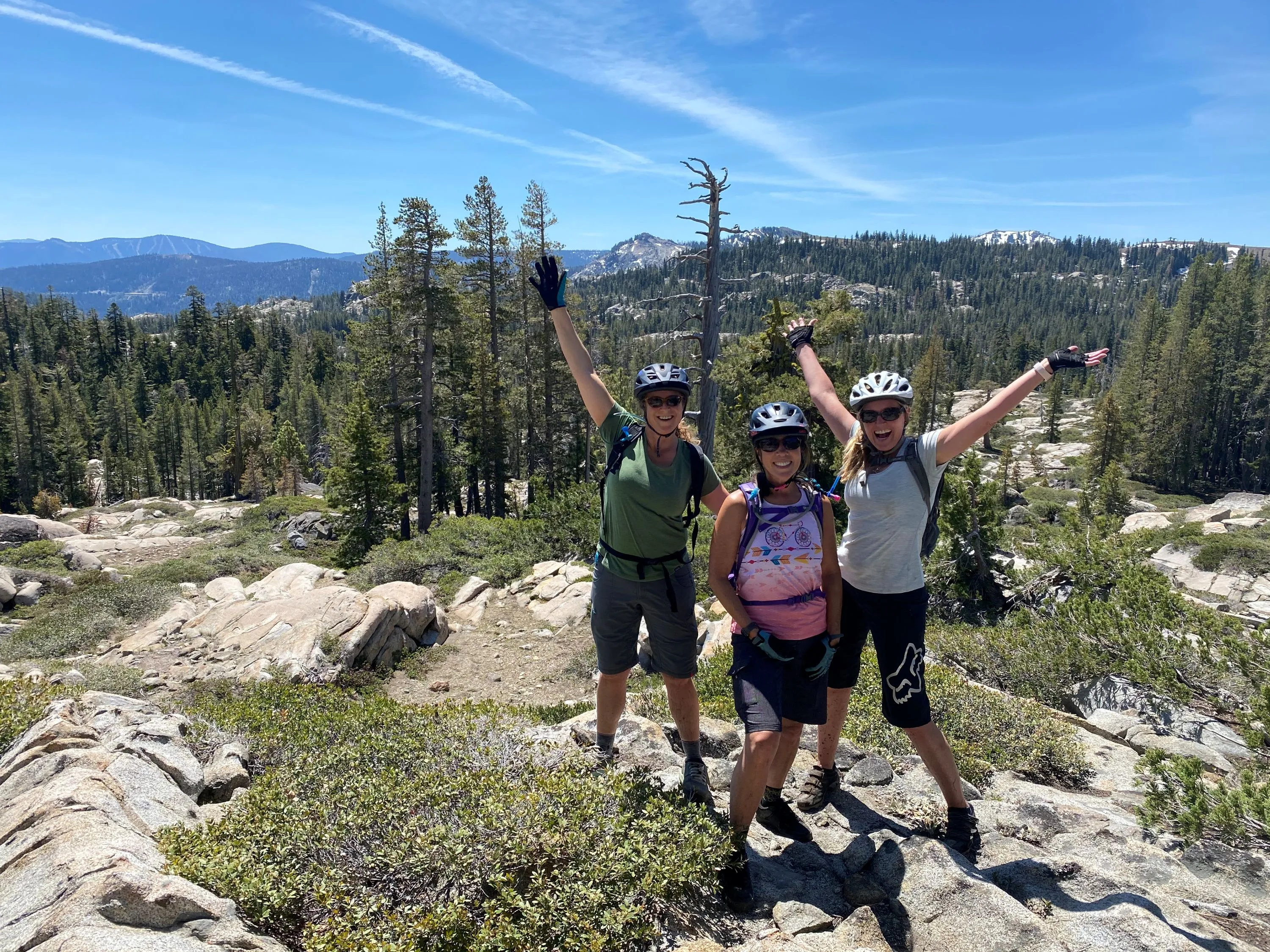 Three hikers wearing helmets and backpacks posing with arms raised on rocky terrain with forested mountains in the background under a clear blue sky.