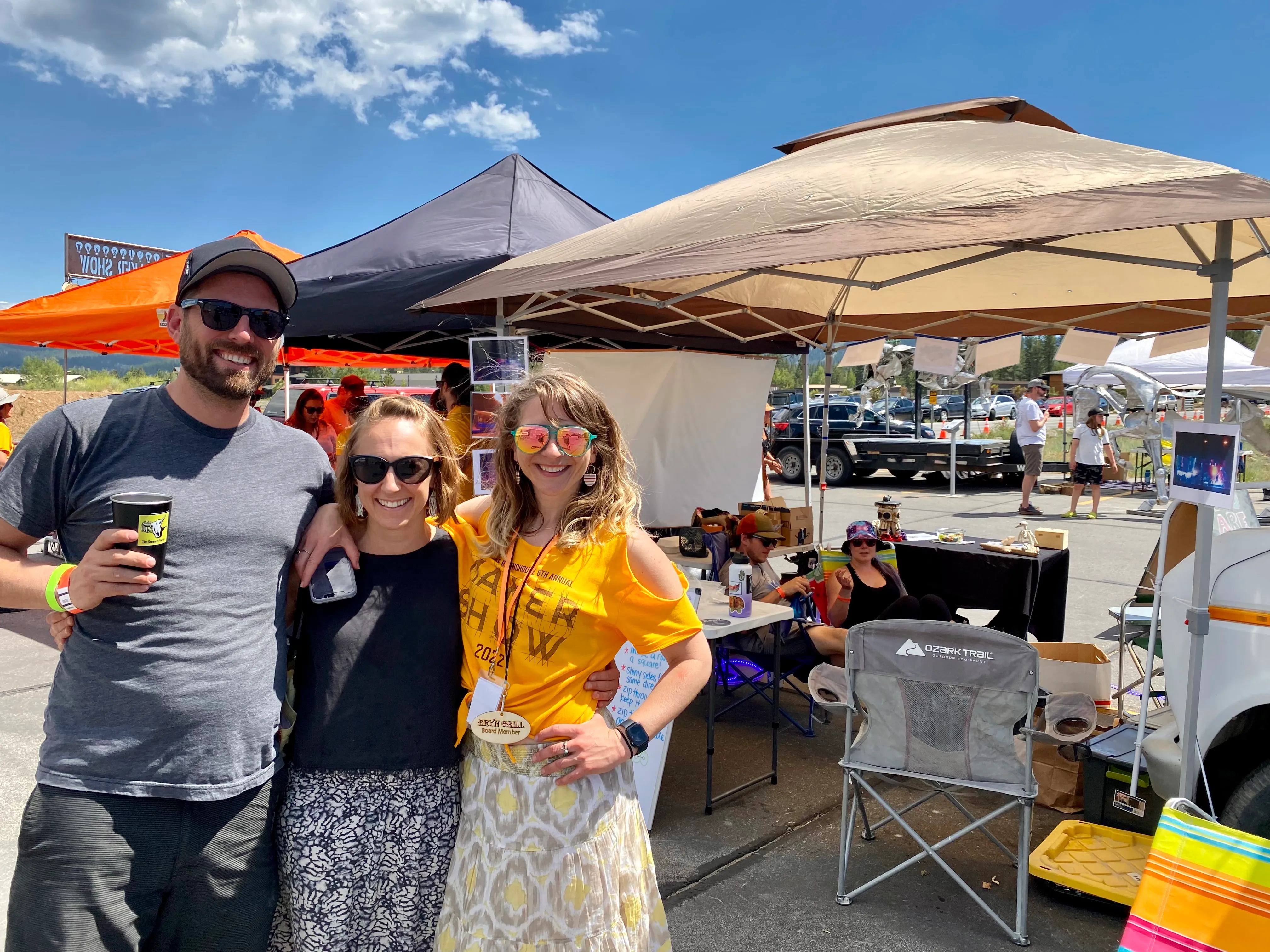 Three smiling people posing outdoors at a market or festival under large canopies on a sunny day.