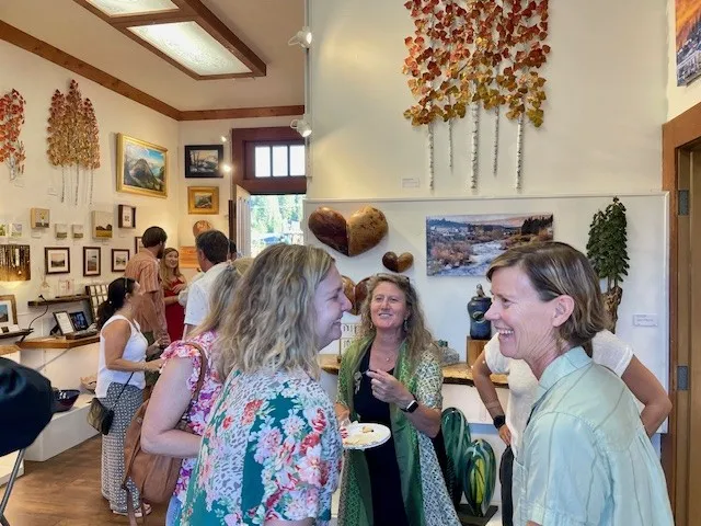 Group of people smiling and socializing inside a warmly lit art gallery with paintings and leaf-themed decorations on the walls.
