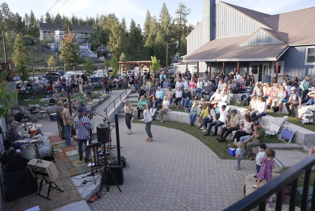 Outdoor concert with a band playing on stage and an audience seated and standing in front, surrounded by trees and buildings.