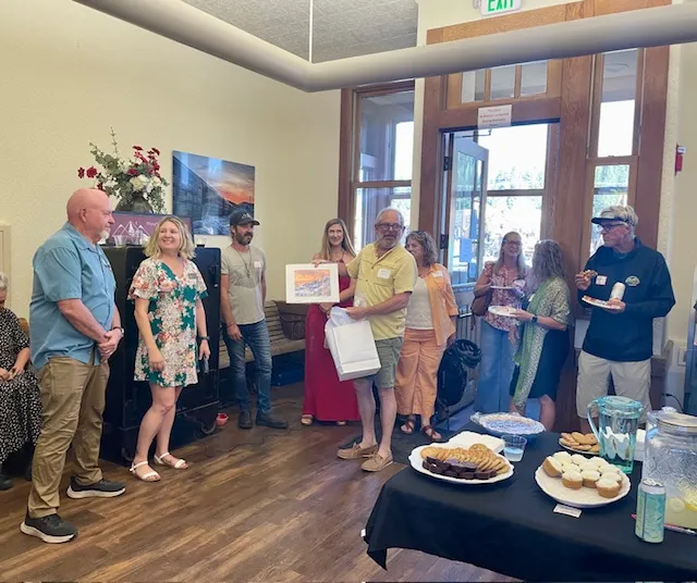 Group of people gathered indoors near a table with desserts, one man holding up a framed picture.
