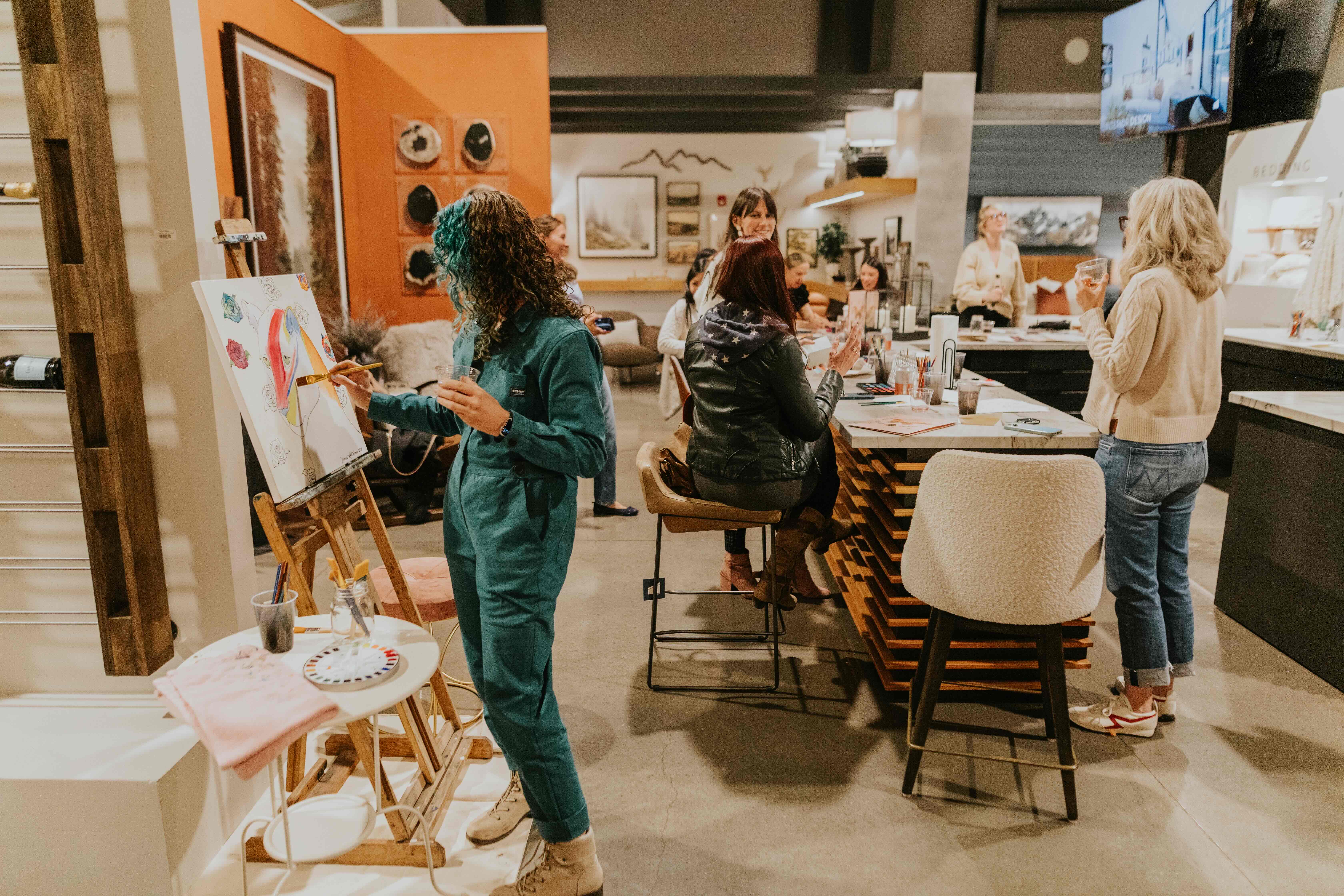 A group of people socializing in a cozy indoor space with one woman painting on a canvas at an easel.