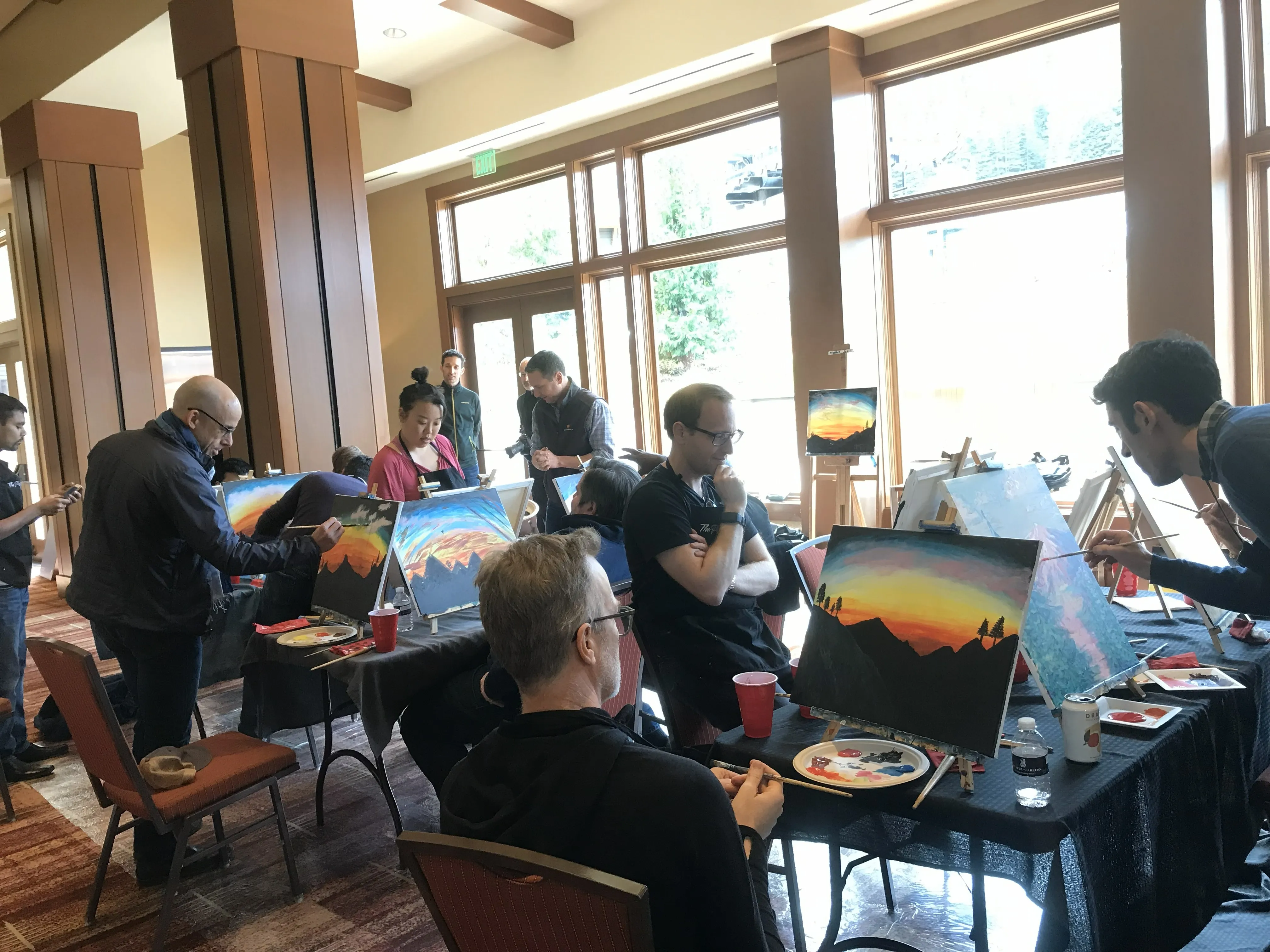 Group of people participating in a painting class indoors, painting sunset landscapes on canvases.