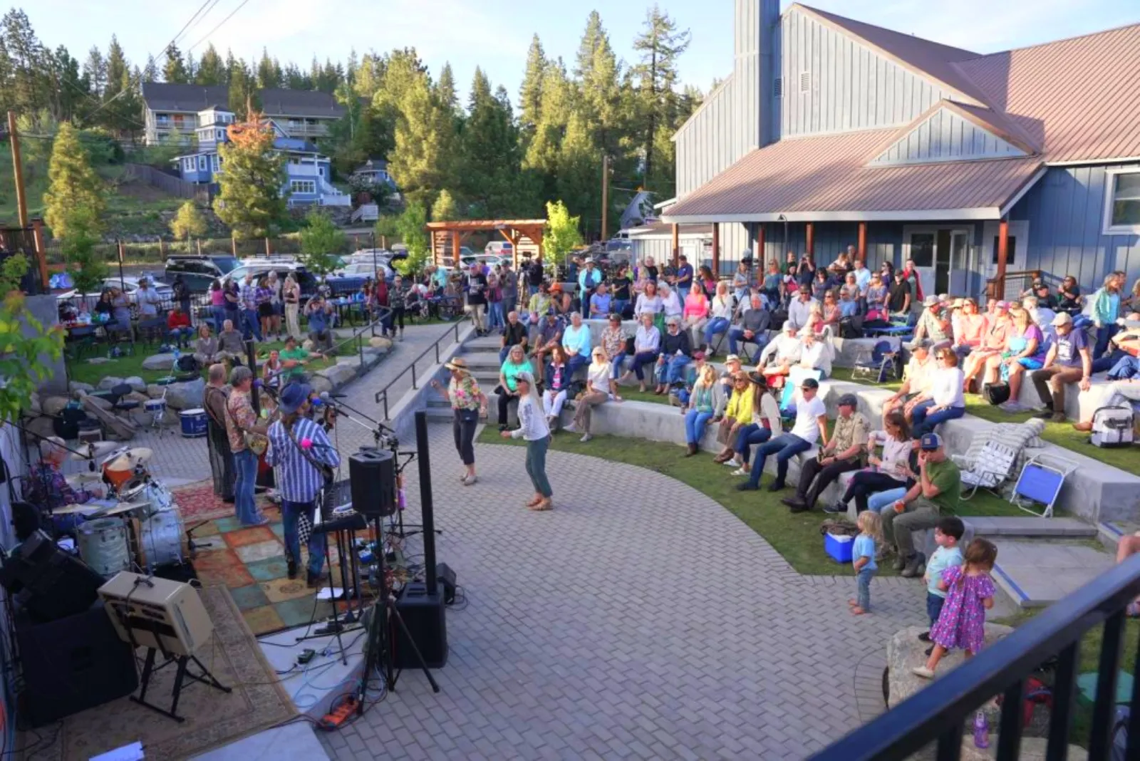 Outdoor community concert with a band playing on stage and a seated audience enjoying the performance in a park-like setting.