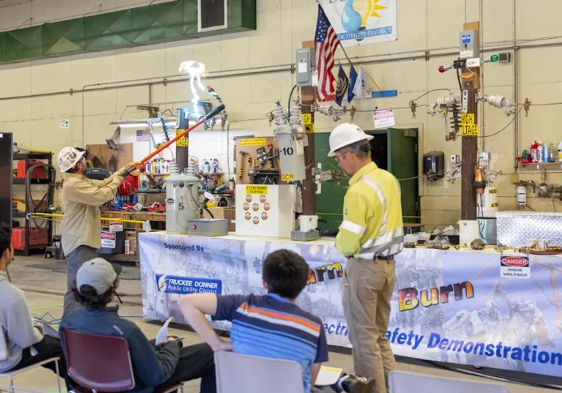 An instructor wearing safety gear demonstrating electrical safety with a safety pole emitting sparks, while an audience watches seated indoors.