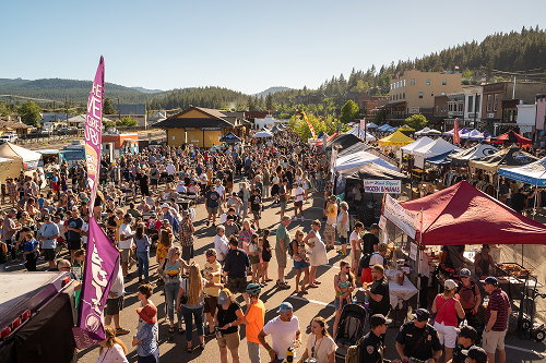 Large crowd of people gathered at an outdoor festival with tents, flags, and a mountainous background under a clear sky.