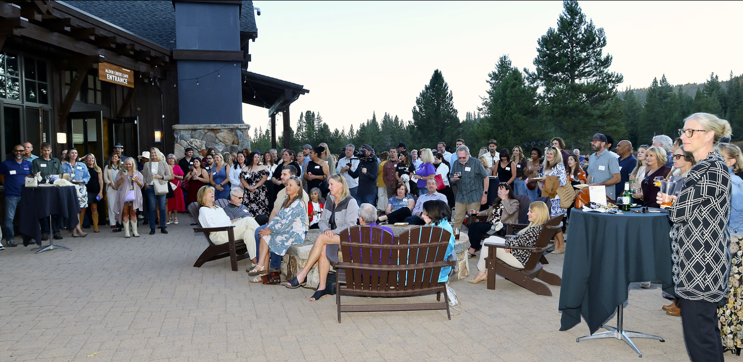 Large group of people socializing and seated around a fire pit outside a building with an 'Alder Creek Café Entrance' sign, surrounded by tall trees.