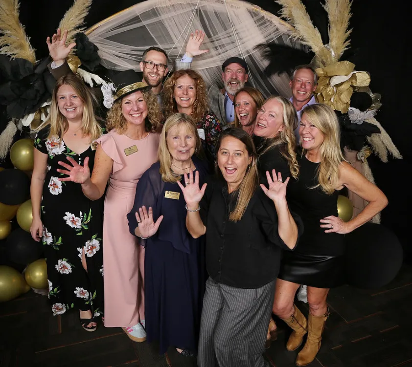 Group of sixteen adults smiling at a party, with many wearing black hats decorated with gold and white feathers, standing in front of black, gold, and white balloons.