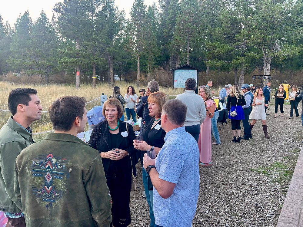 Large group of people socializing and seated around a fire pit outside a building with an 'Alder Creek Café Entrance' sign, surrounded by tall trees.