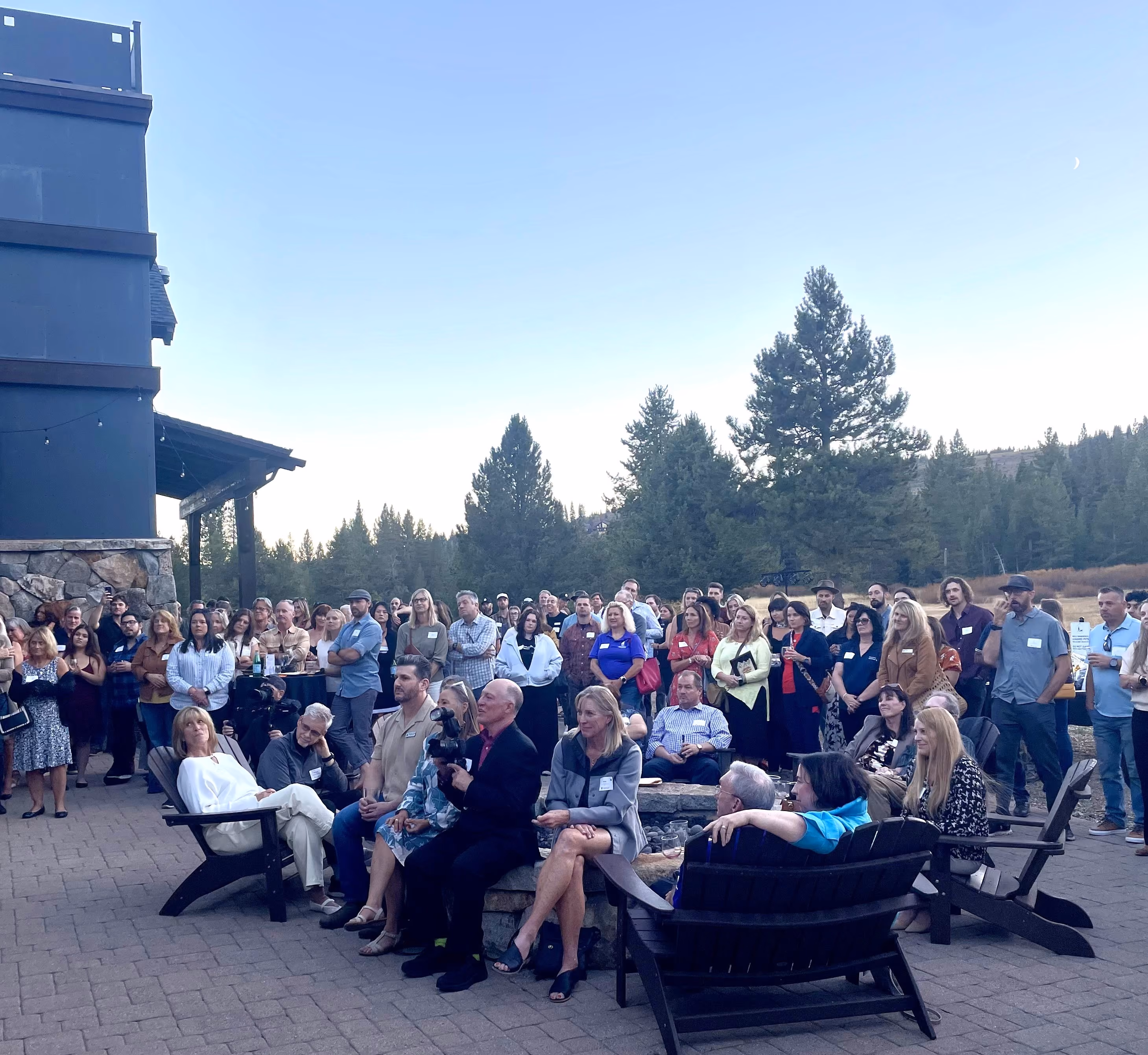 Large group of people socializing and seated around a fire pit outside a building with an 'Alder Creek Café Entrance' sign, surrounded by tall trees.