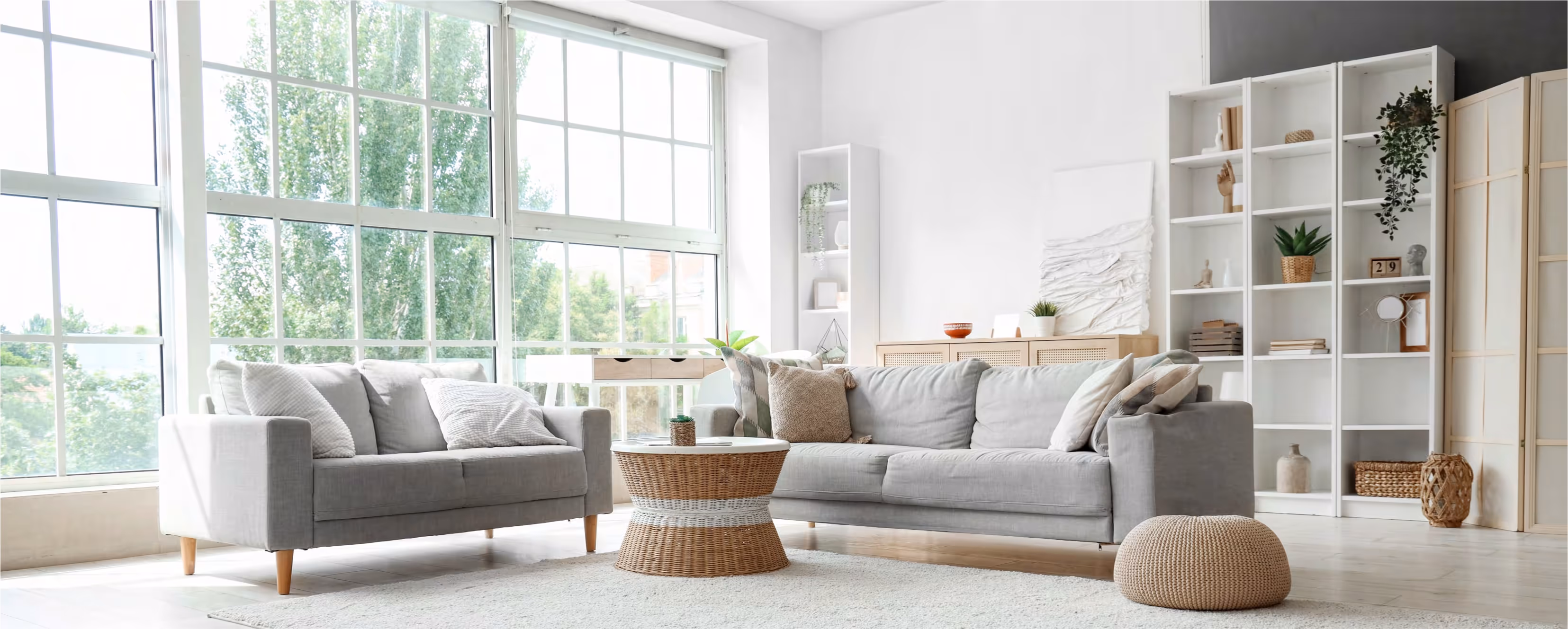 Bright living room with large windows, two gray sofas with pillows, a wicker coffee table, and white shelving with decor.