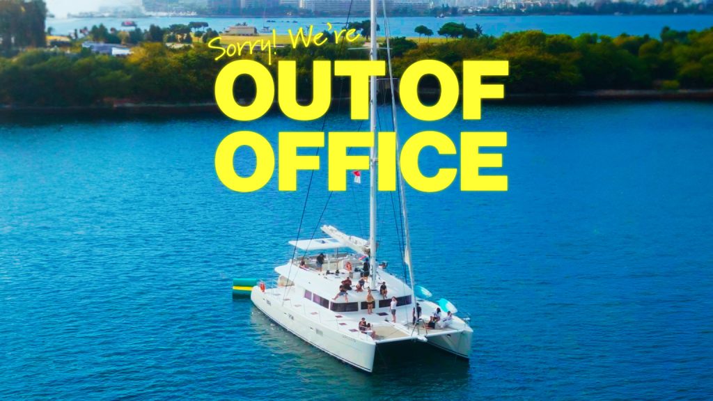 White catamaran sailboat in blue water with "Sorry We're Out of Office" text overlay