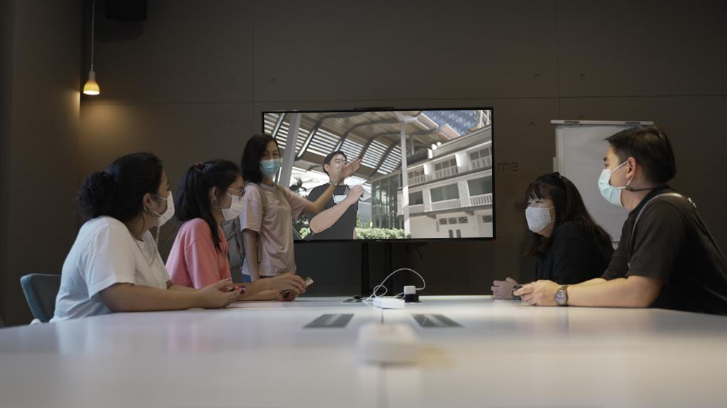 Group of masked people in video conference meeting around table with large screen