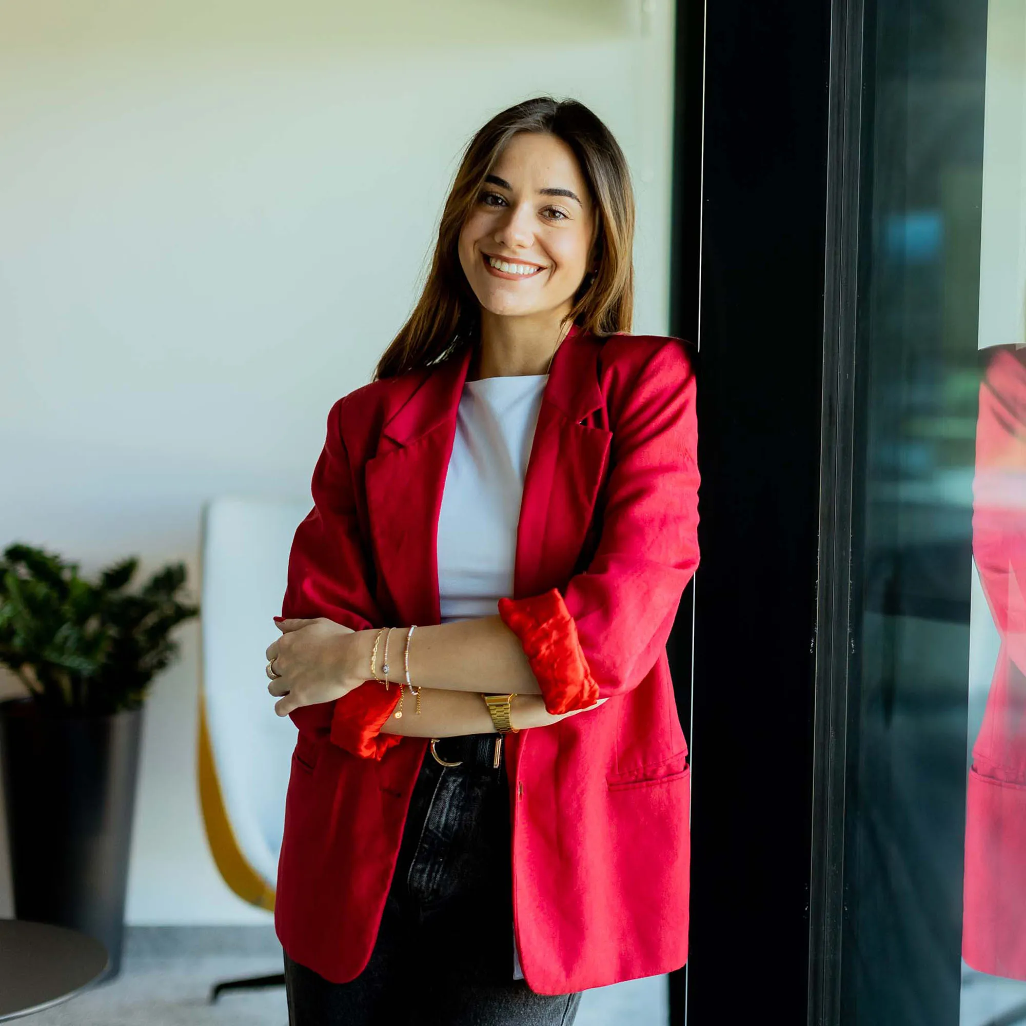 Smiling woman in a red blazer leaning against a glass wall in an office.