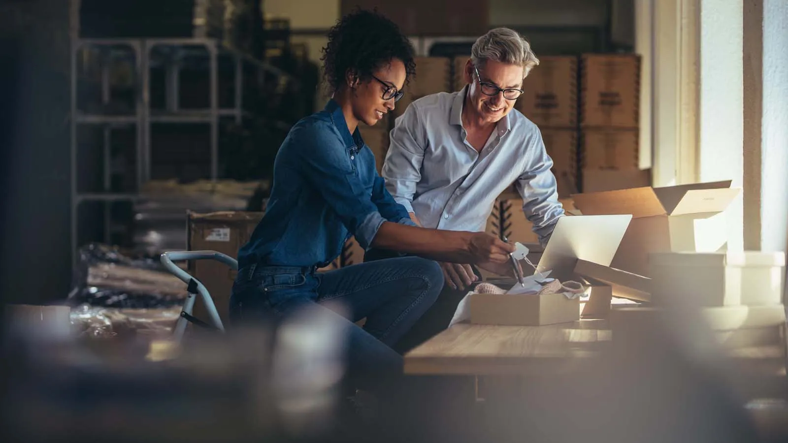Two people in a warehouse packing a box while looking at a laptop together.