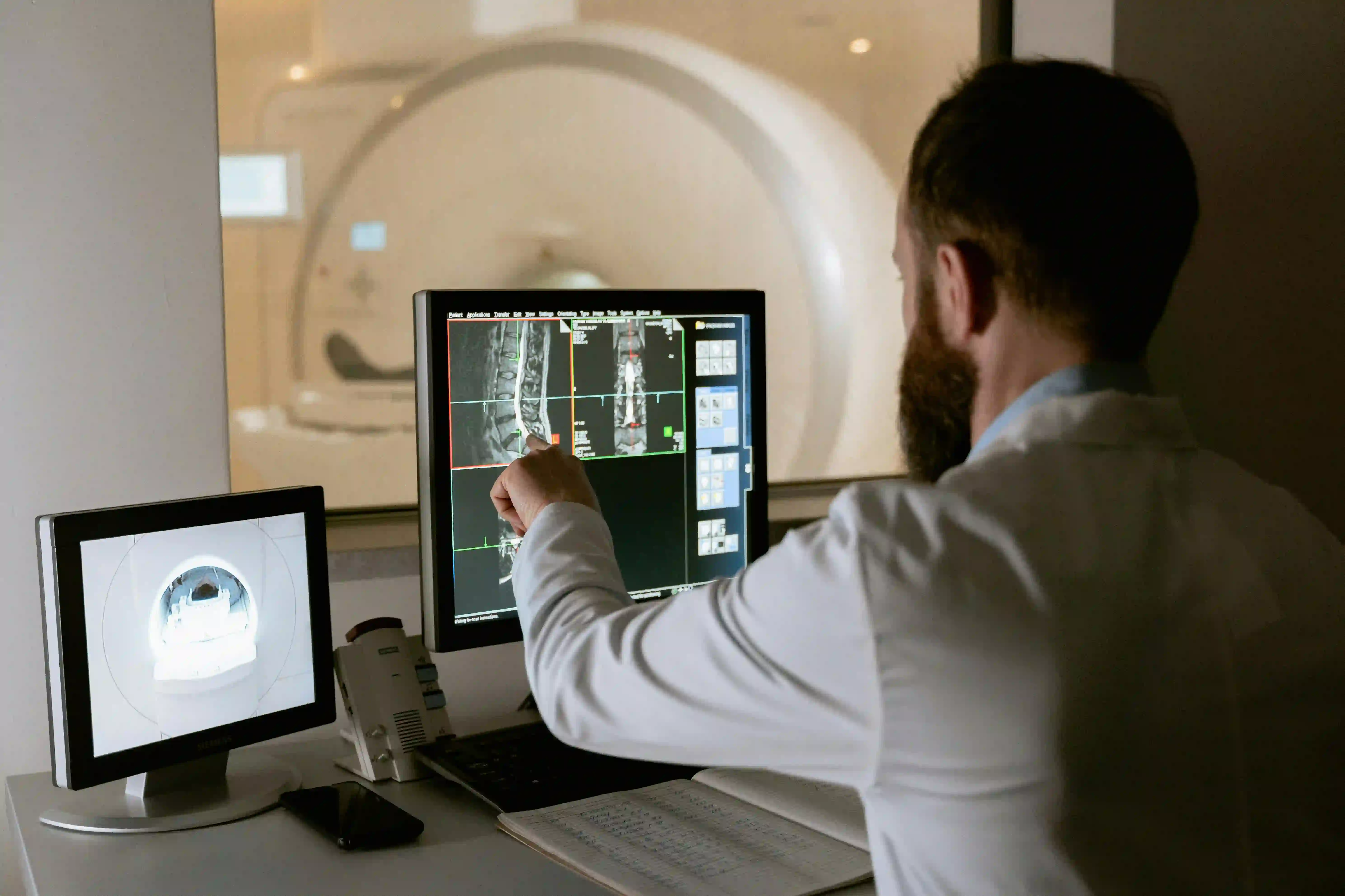 Doctor in white coat analyzing spinal MRI scans on a computer screen next to an MRI machine visible through a window.