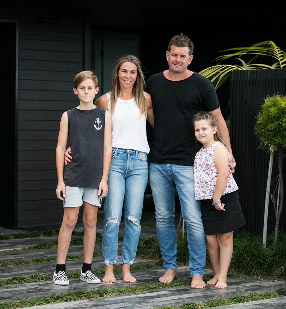 A barefoot family of four standing outdoors on a stone path with green grass, posing in front of a modern dark building.