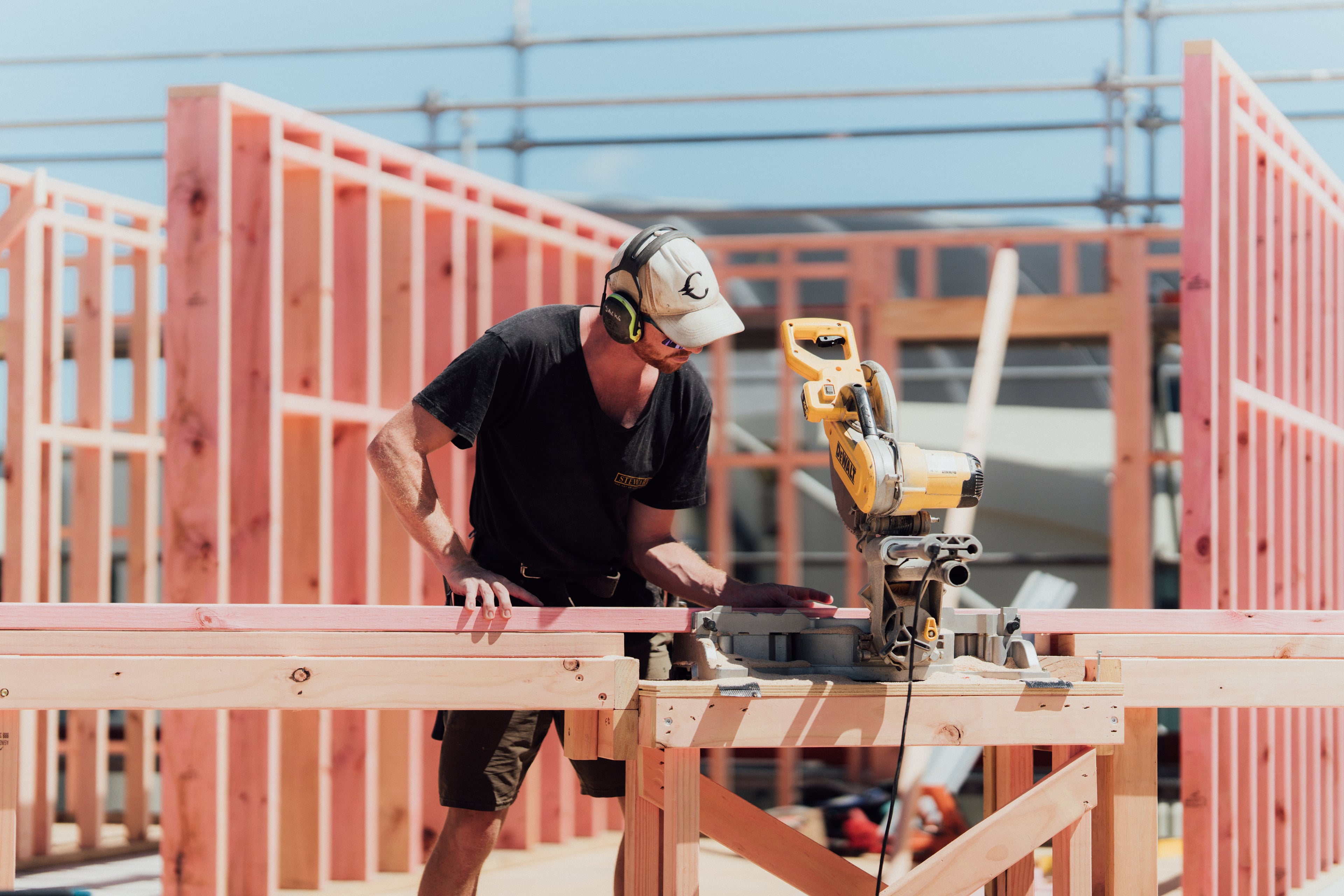 Construction worker wearing ear protection and cap operating a power saw to cut wooden planks at a building site.