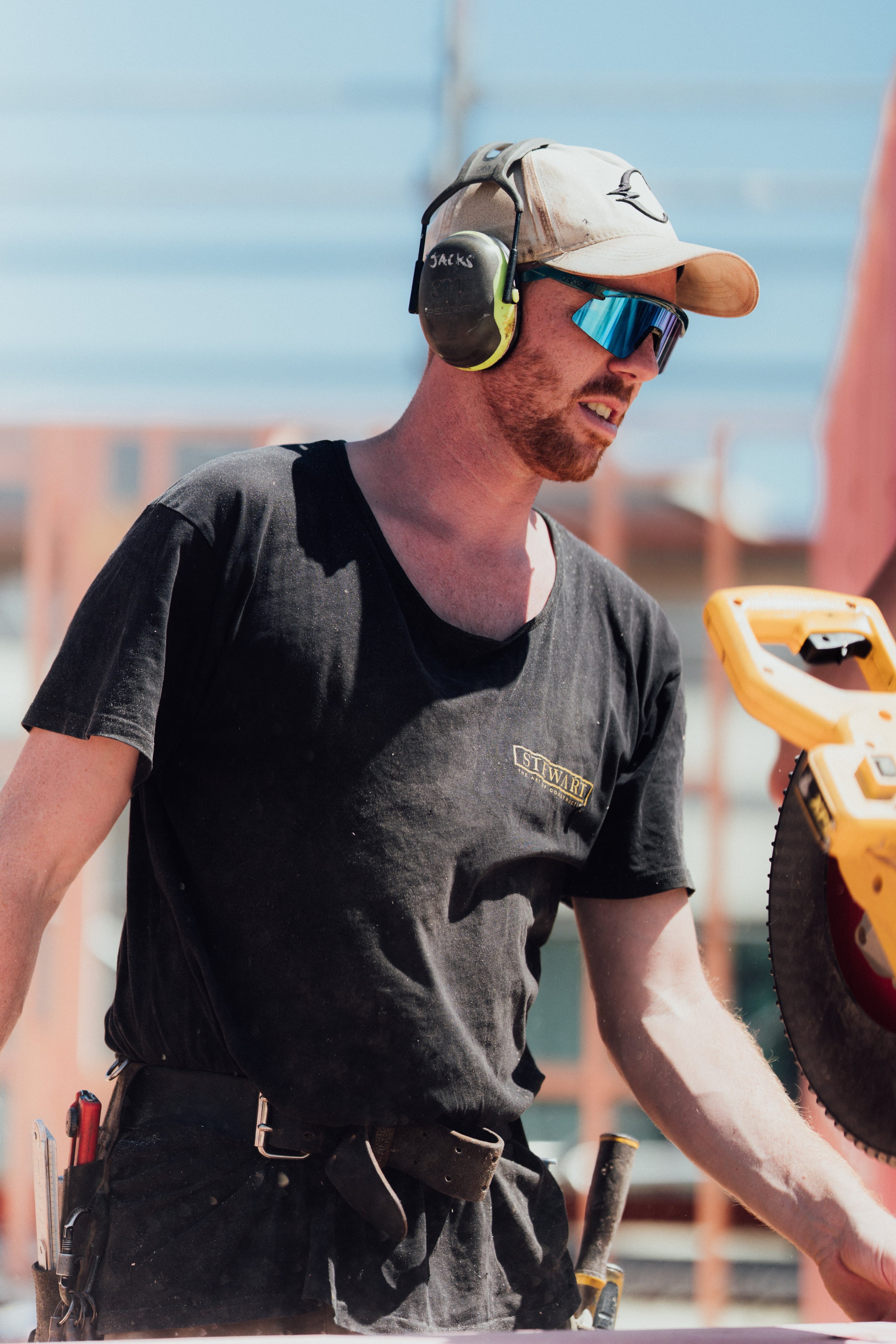 Construction worker wearing a black Stewart Construction shirt, safety earmuffs, a beige cap, and blue reflective sunglasses operating a yellow circular saw outdoors.