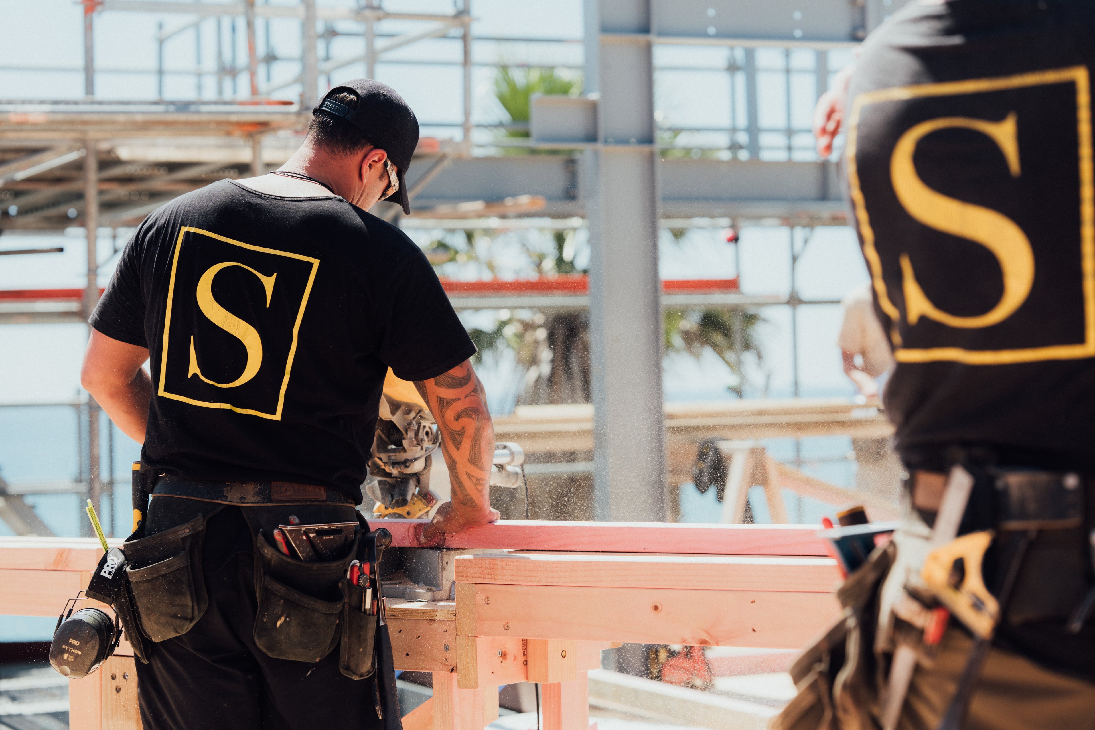 Two construction workers with tool belts working on wooden beams, one using a power saw.