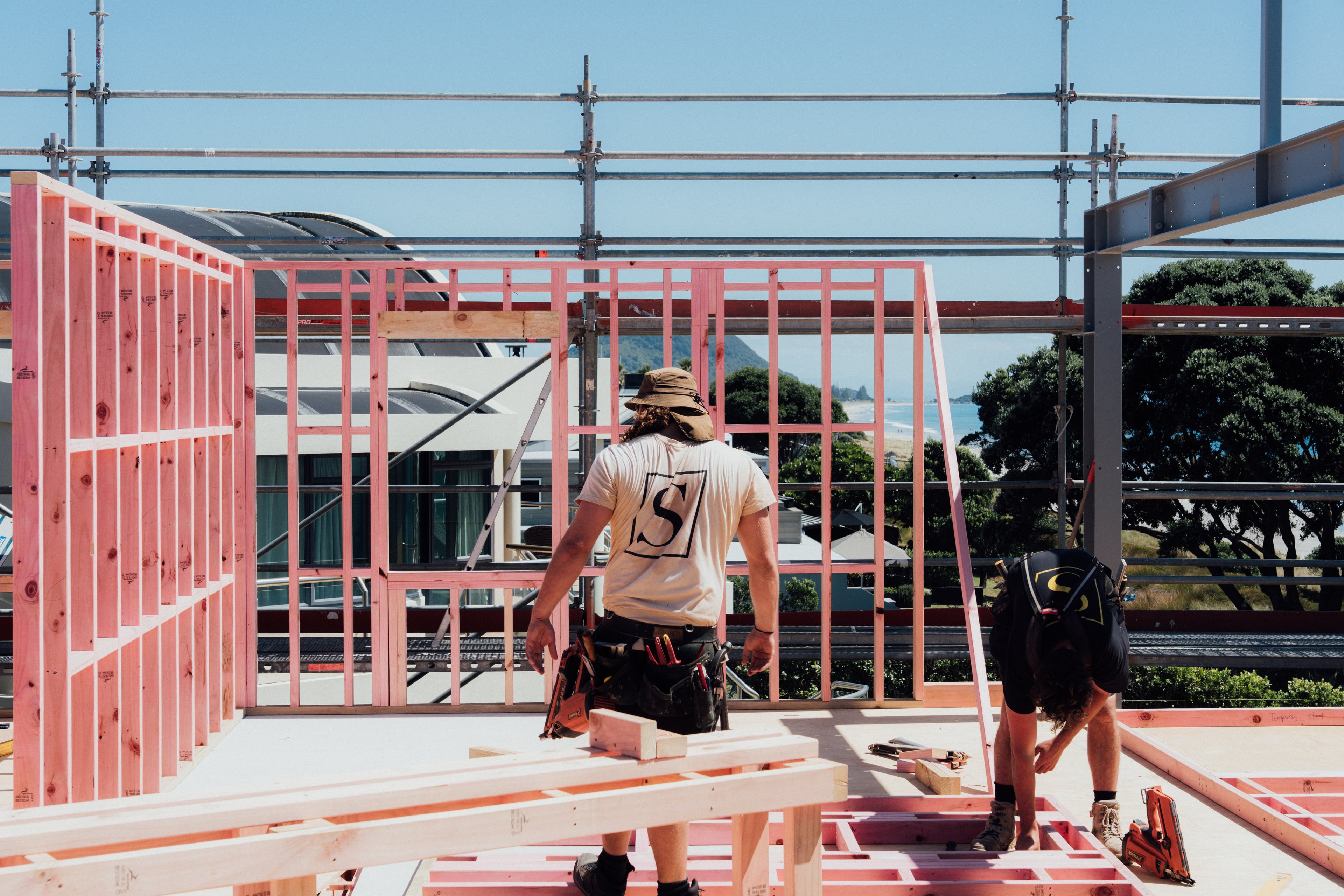 Two construction workers framing walls with pink timber on a building site near a coastline.
