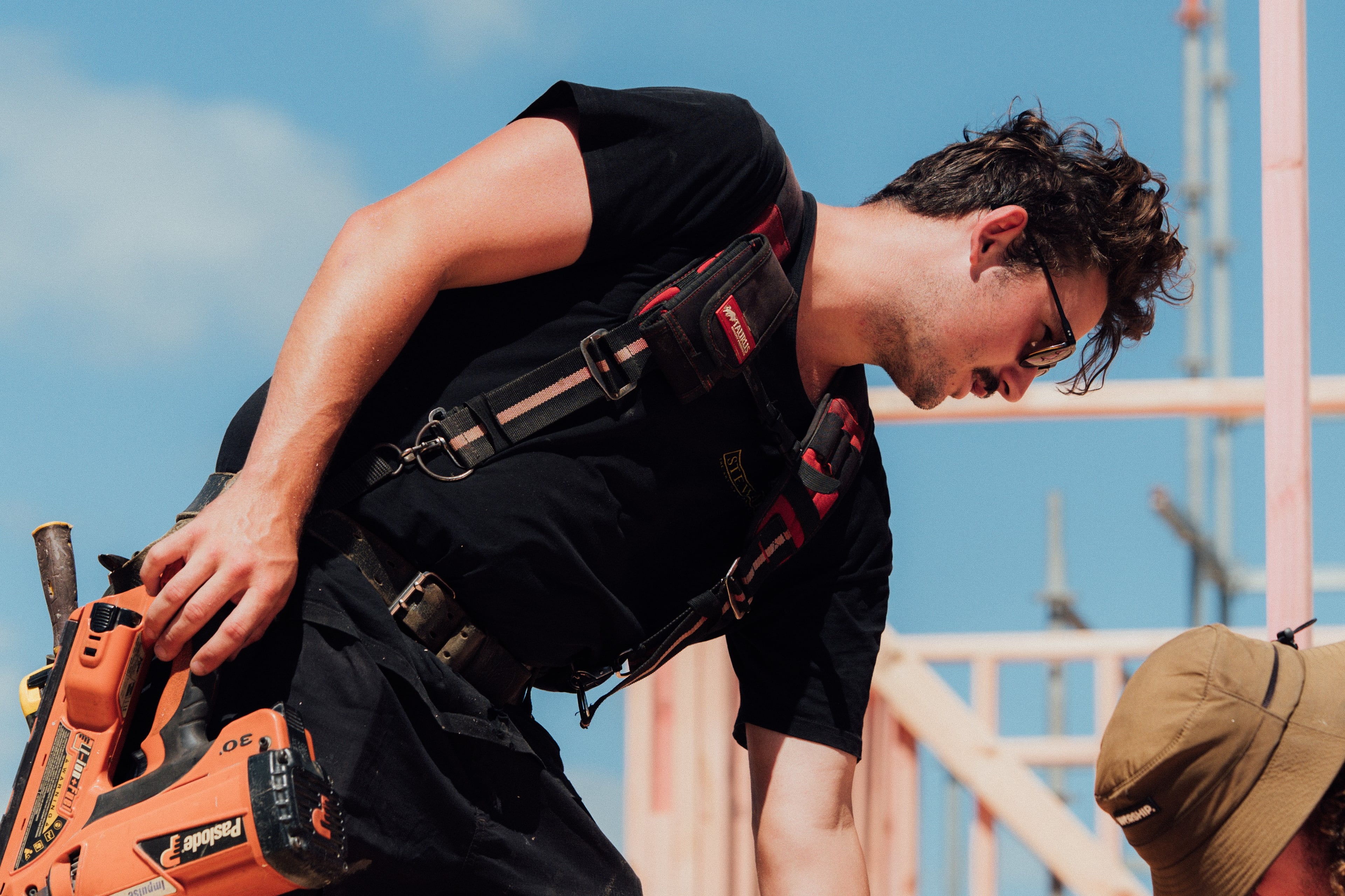 Construction worker wearing black shirt and safety harness holding an orange nail gun on a building site.