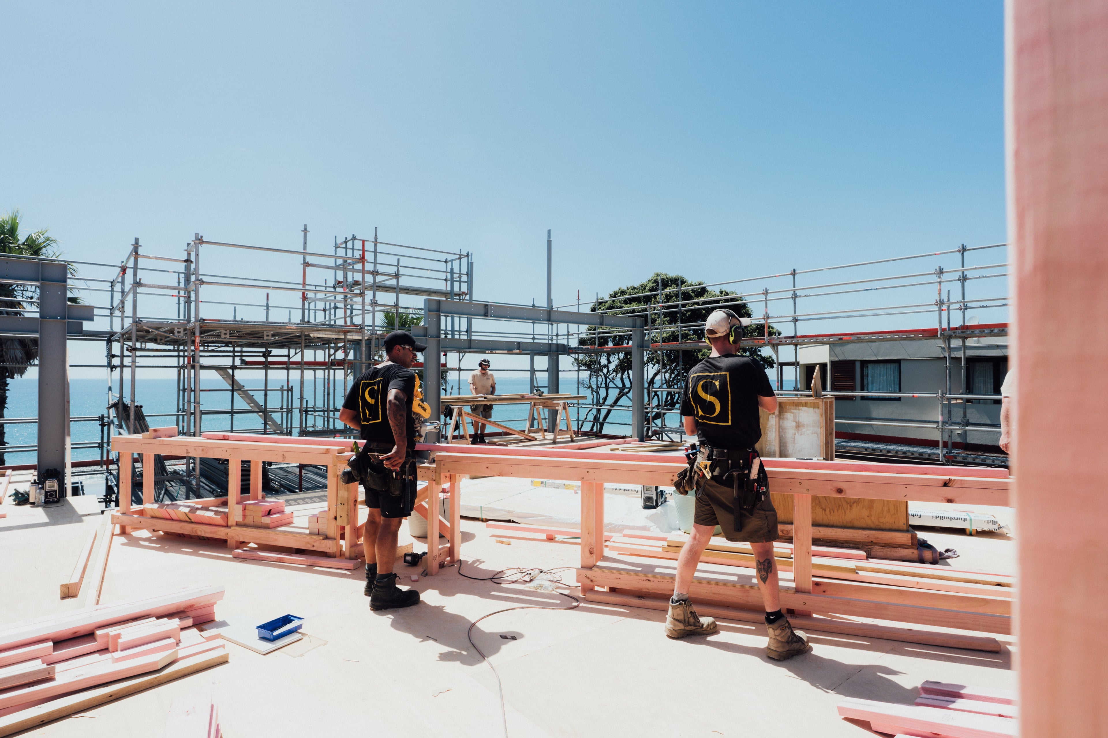 Two construction workers handling wooden beams on a building site with scaffolding and ocean view.