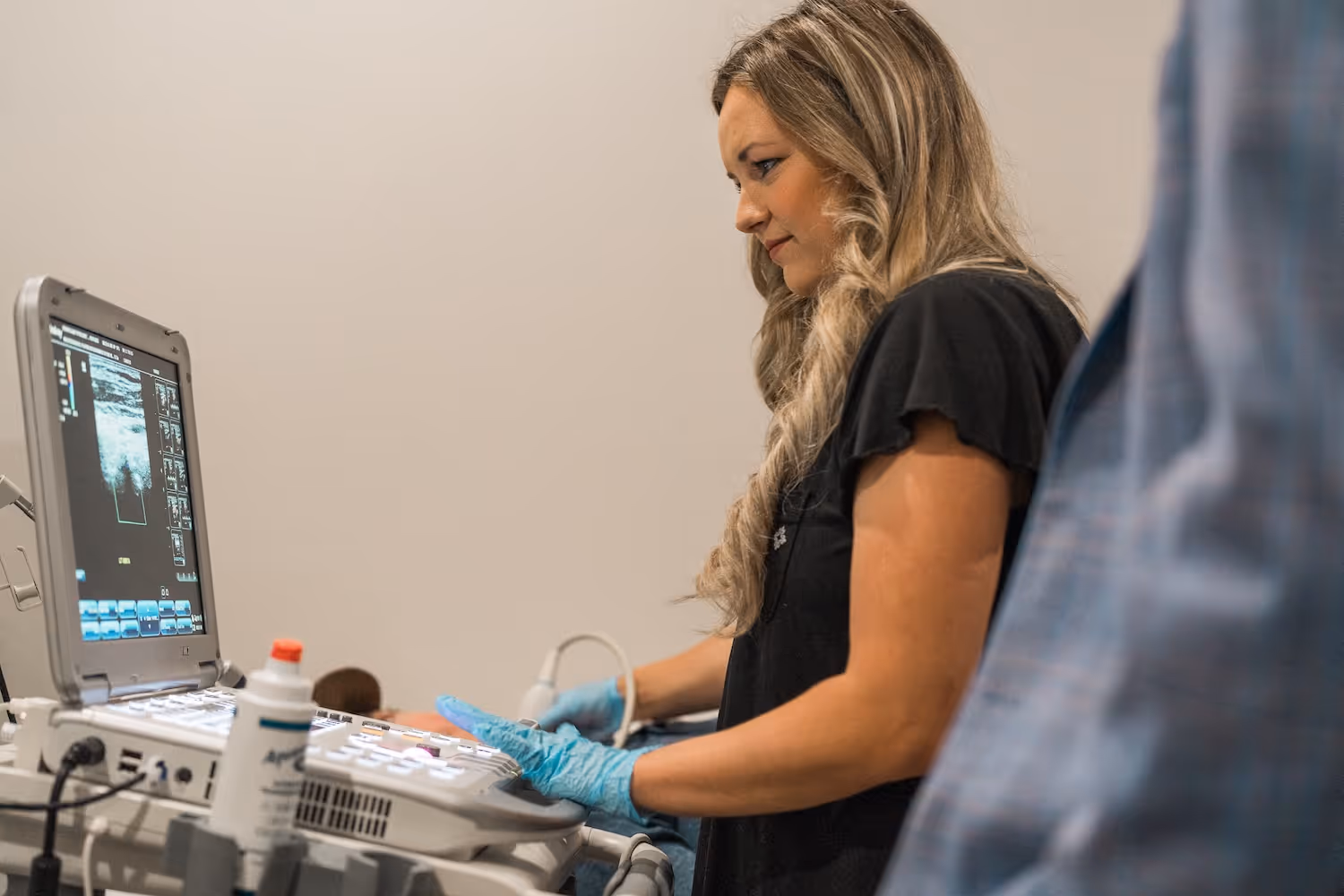 Woman wearing blue gloves operating an ultrasound machine with the ultrasound image visible on the screen.