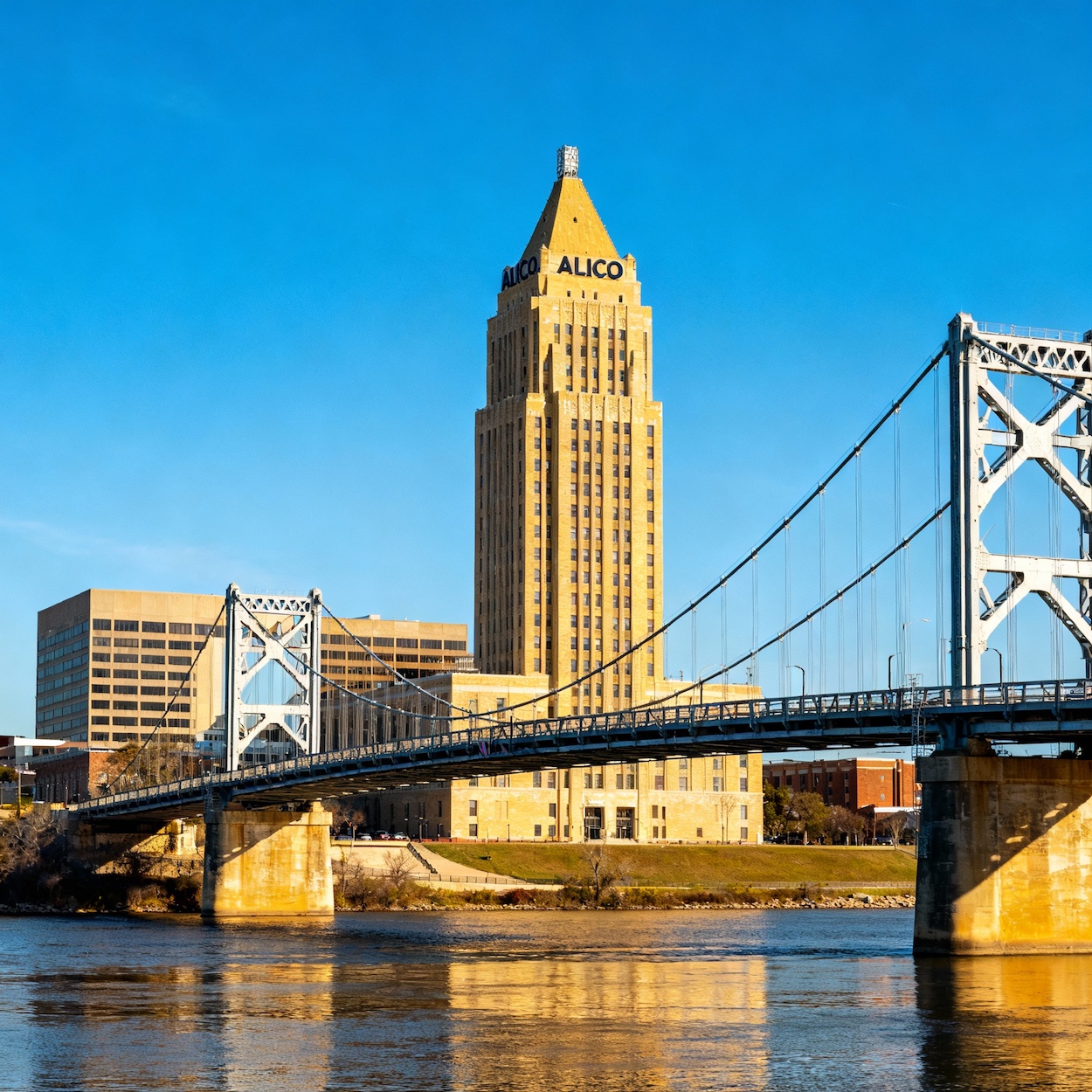 Photograph-style image of the Waco Suspension Bridge spanning the Brazos River in Waco, Texas, showing the bridge's cable suspension structure with calm river water below and a clear sky, a noted historic landmark in downtown Waco.