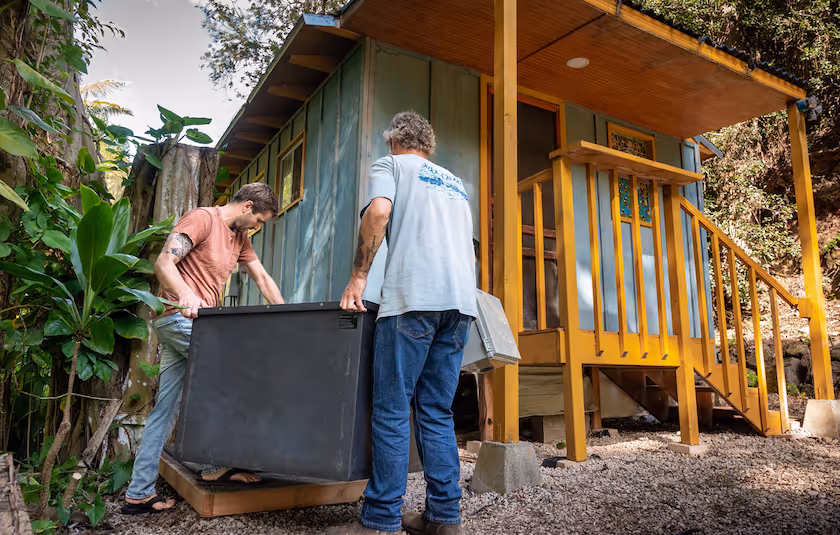 Two men carrying a Mata to install it next to a hawaiian hale, the home is post and pier.