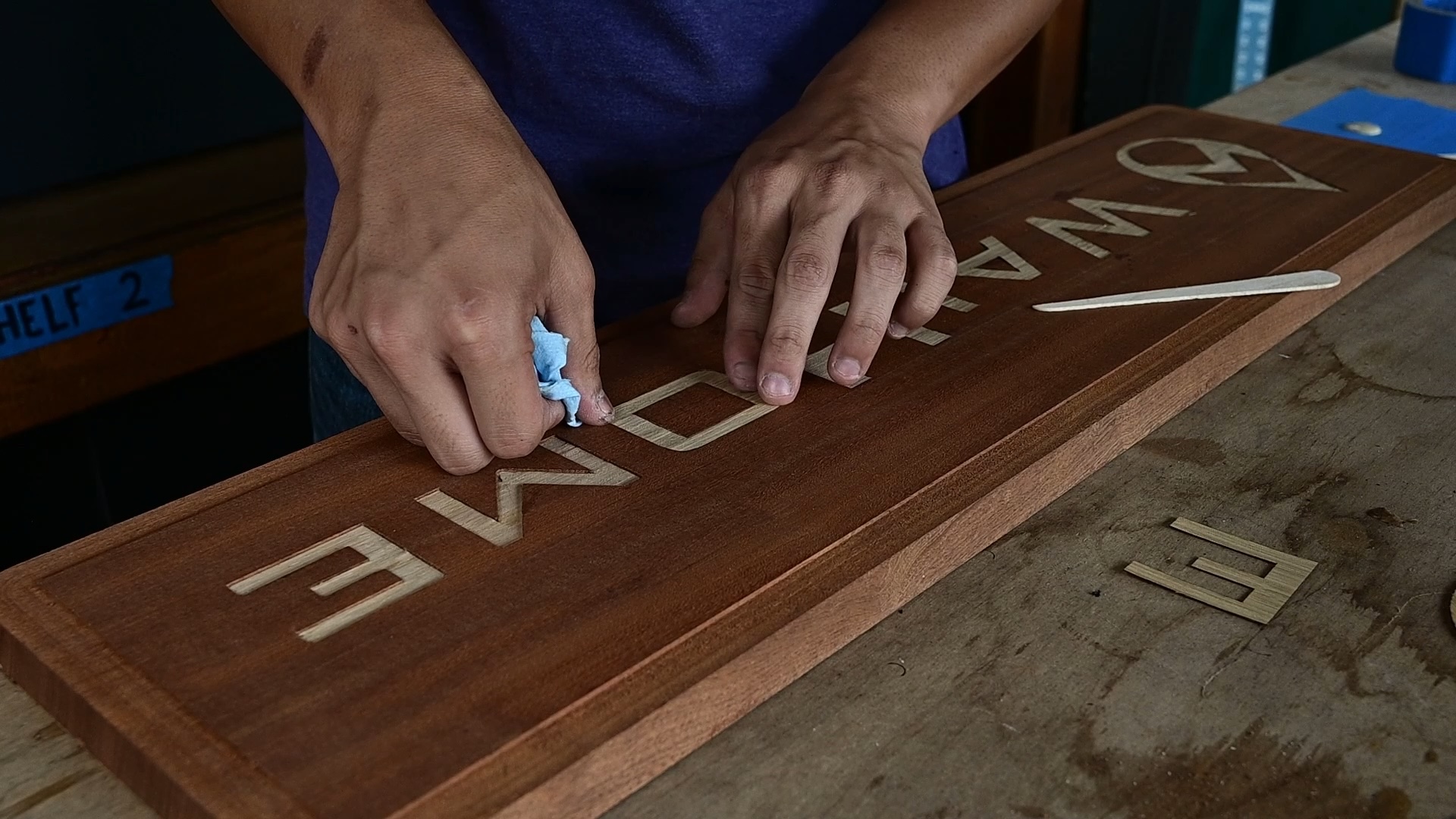 Close-up of a person using a blue cloth to polish carved letters on a wooden sign that reads 'AWANE' placed on a workbench.