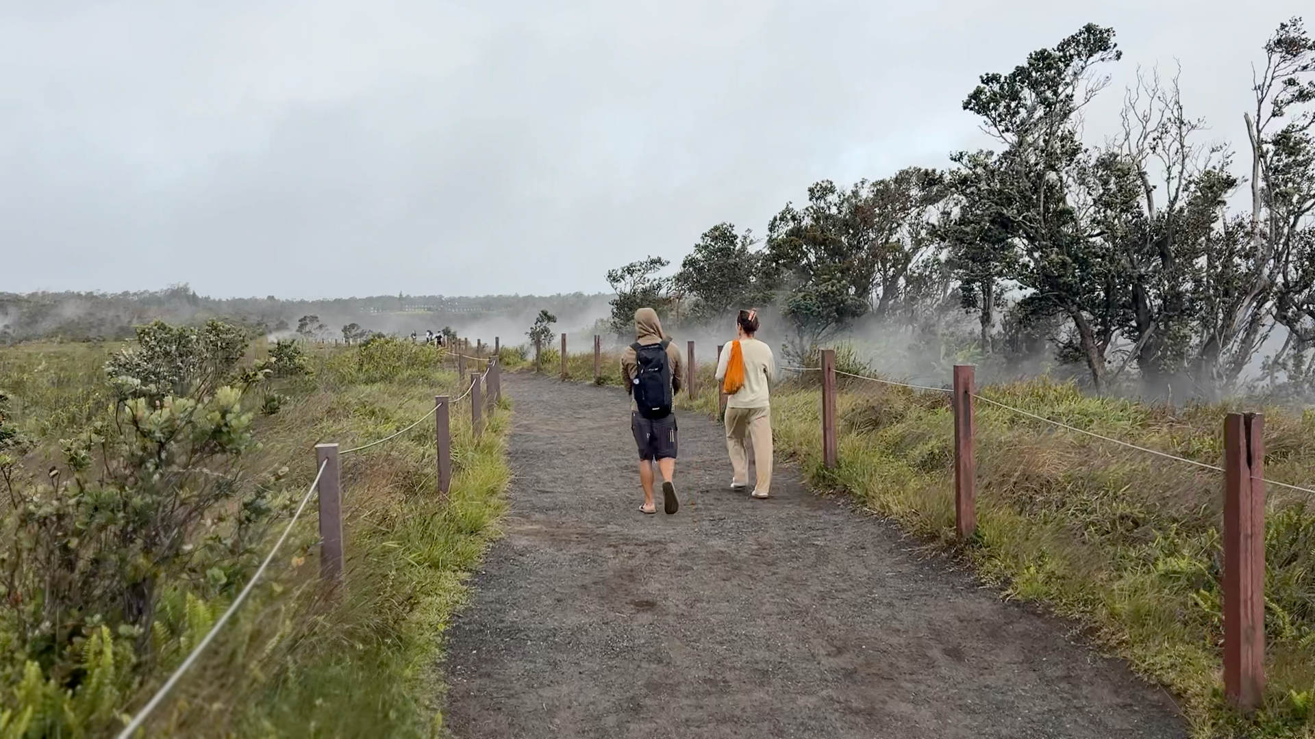 Greg and Natasha walking on a dirt path bordered by wooden posts and rope with grass and trees on either side, at the Kilauea volcano.