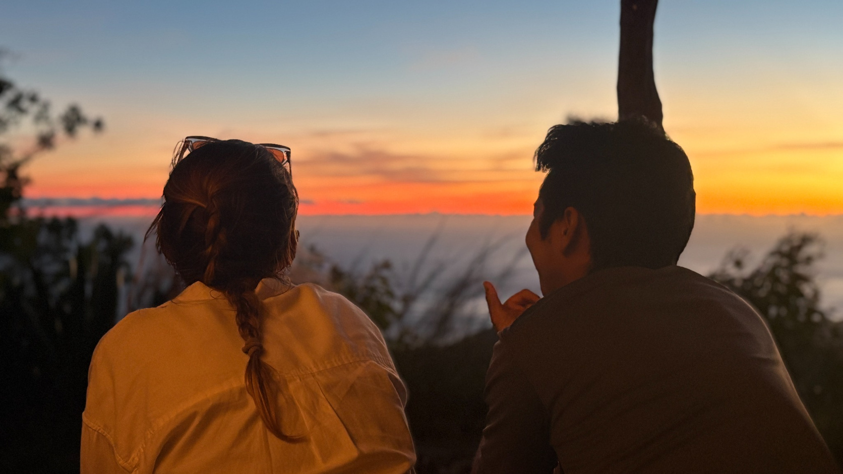 Two people sitting outdoors facing a vibrant sunset with orange, pink, and blue hues in the sky, one person with braided hair and glasses resting on their head, the other gesturing with their right hand.