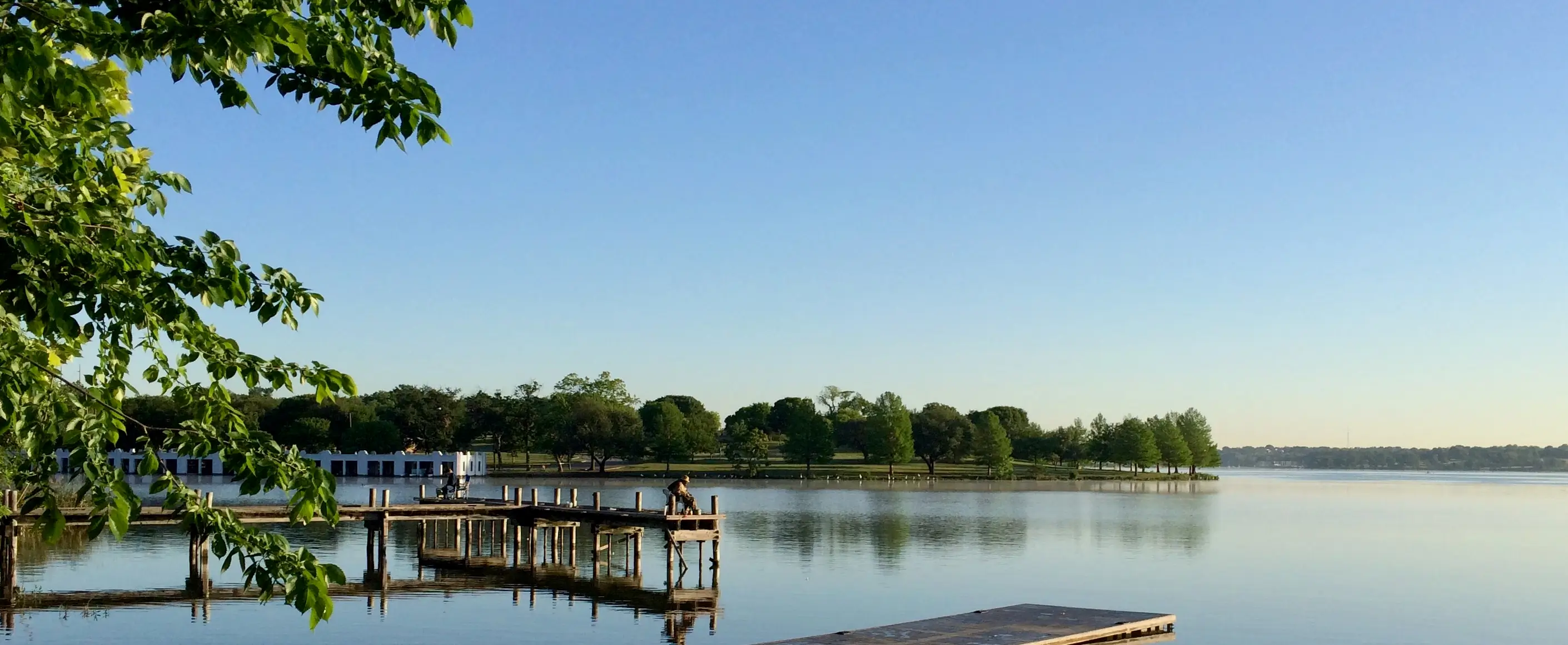 Person fishing at Dallas White Rock Lake a wooden pier extending into a calm lake with trees and a building in the background under a clear sky.
