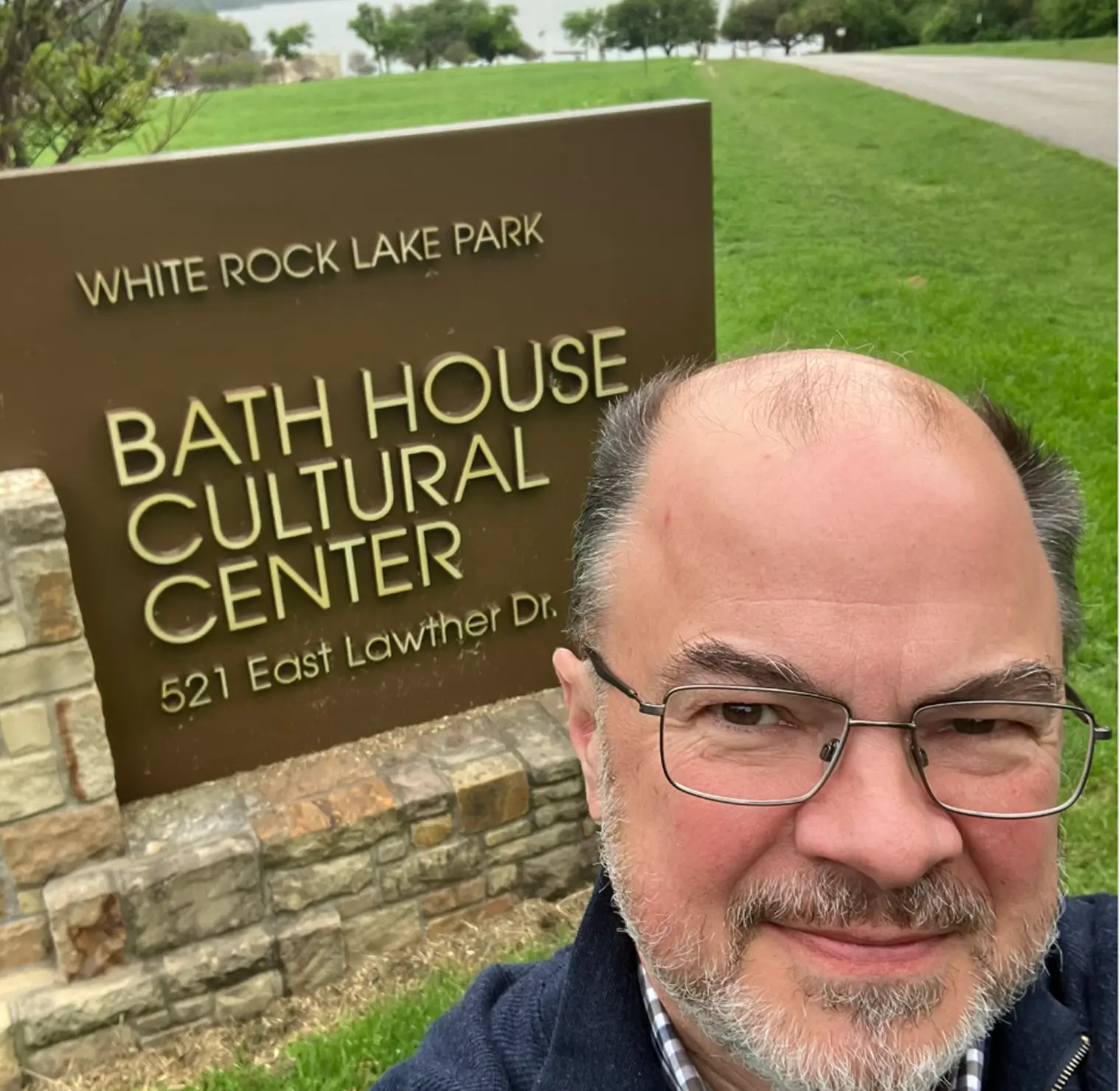 Man with glasses and a beard taking a selfie in front of a stone and metal sign that reads 'White Rock Lake Park Bath House Cultural Center 521 East Lawther Dr.'