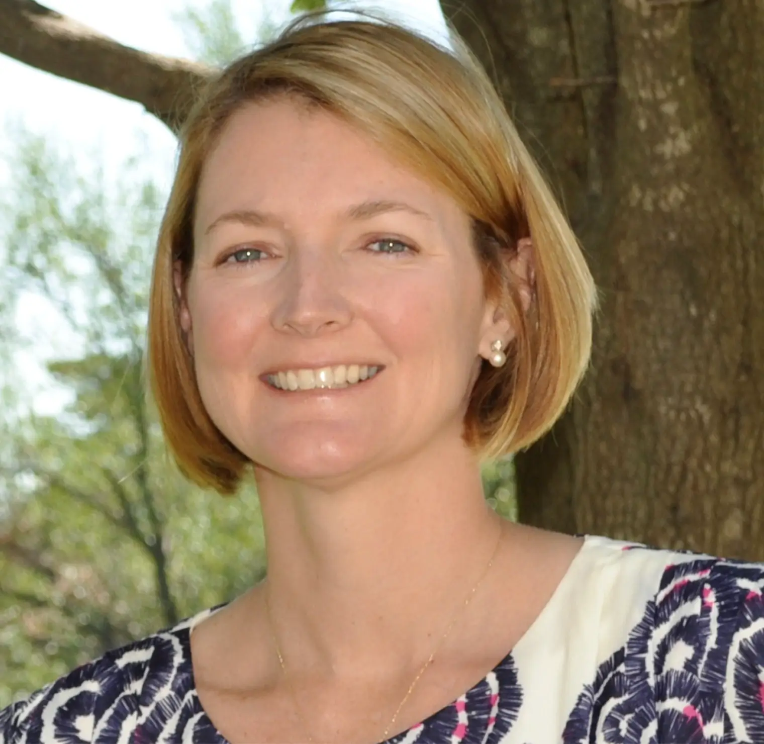 Smiling woman with short blonde hair wearing pearl earrings and patterned top outdoors near a tree.