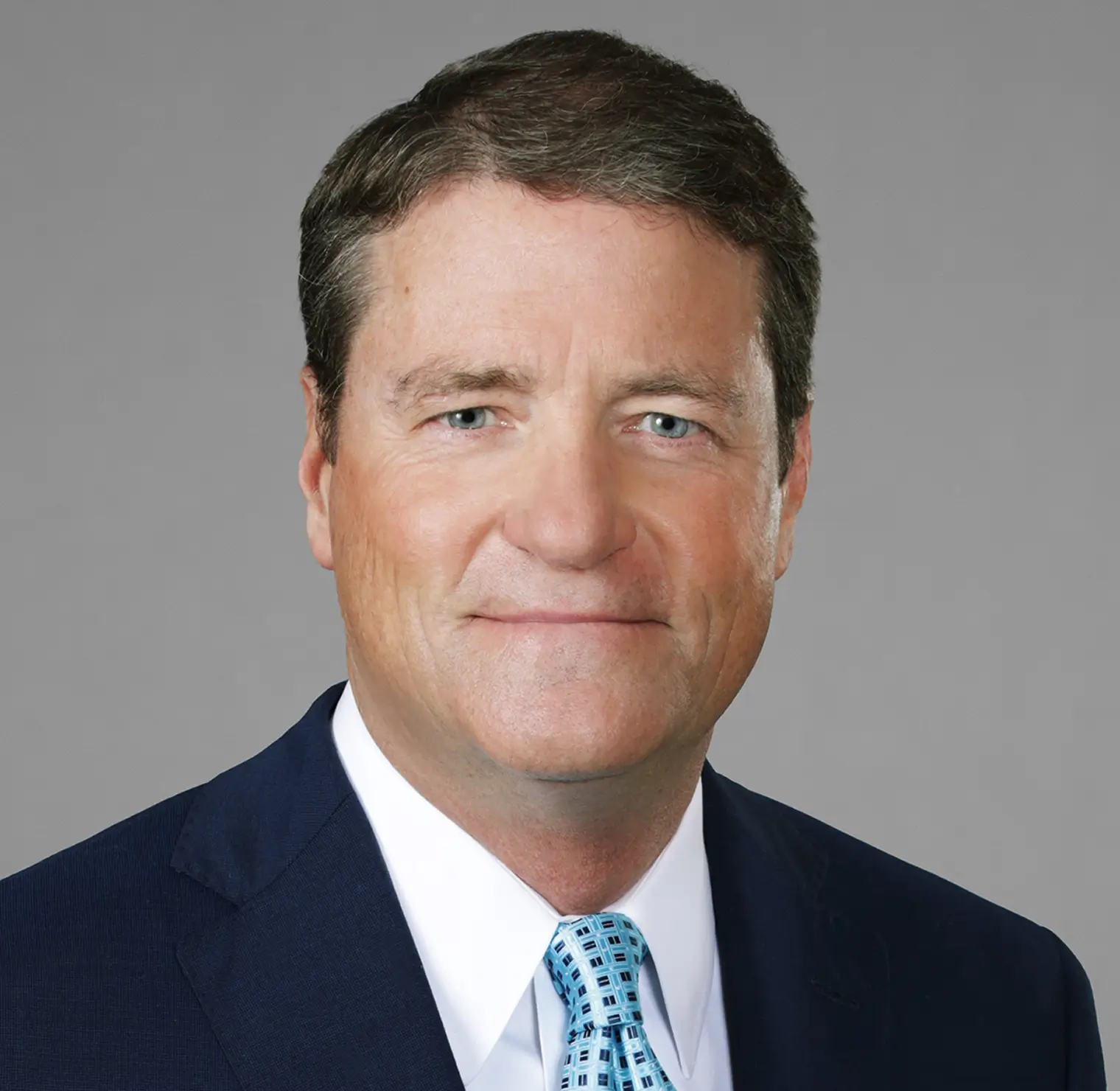 Headshot of a middle-aged man with blue eyes and brown hair wearing a dark blue suit and light blue patterned tie.