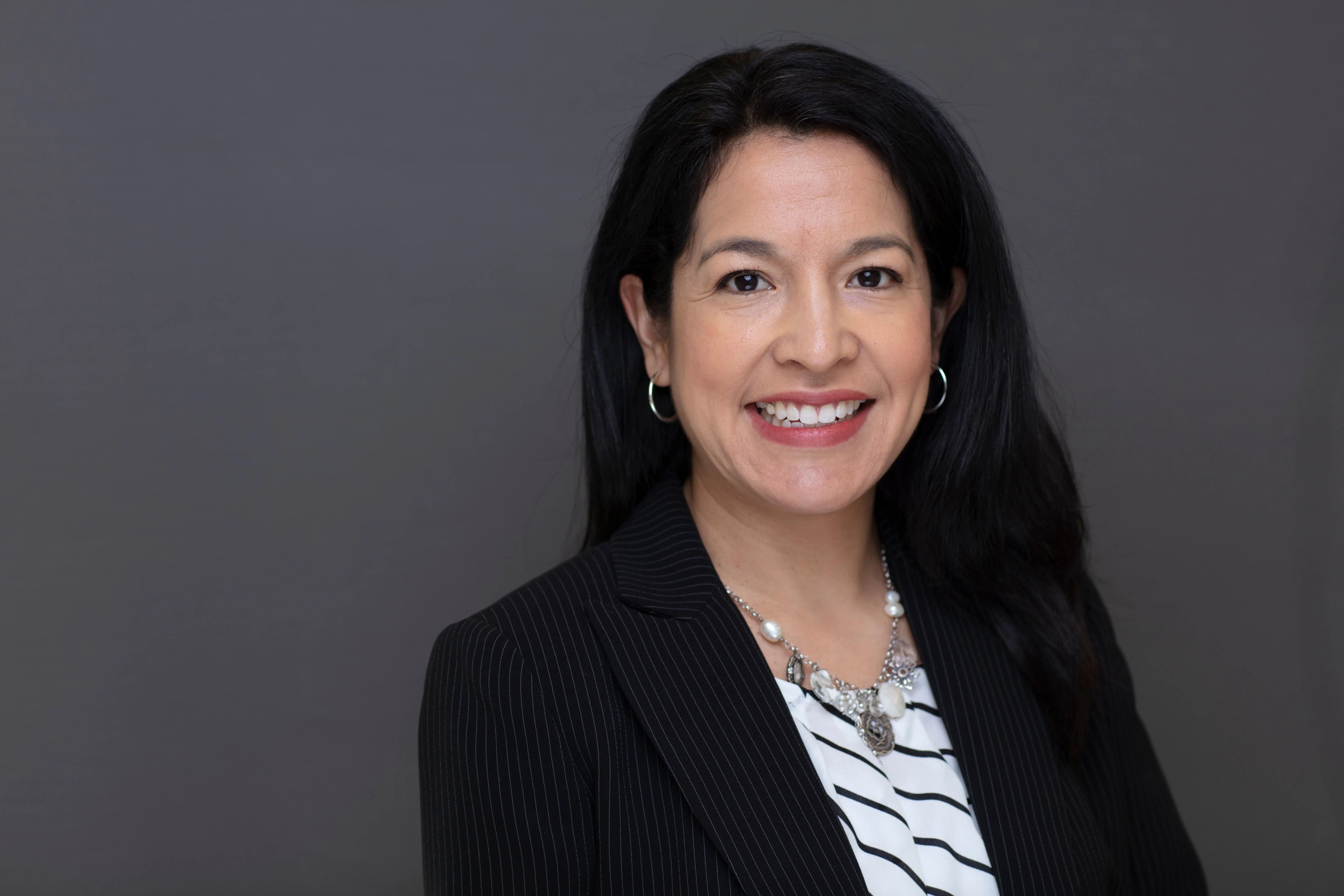 Smiling woman with long black hair wearing hoop earrings, a striped black blazer, and a striped white blouse with a necklace against a gray background.