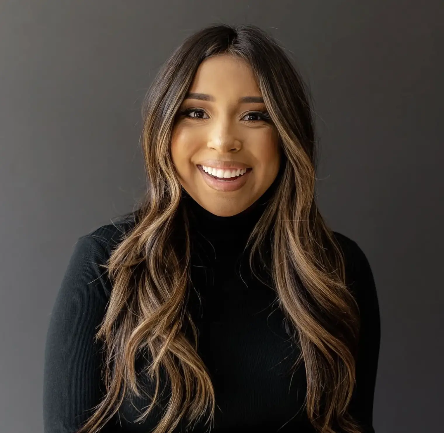 Smiling woman with long brown hair wearing a black turtleneck against a dark gray background.