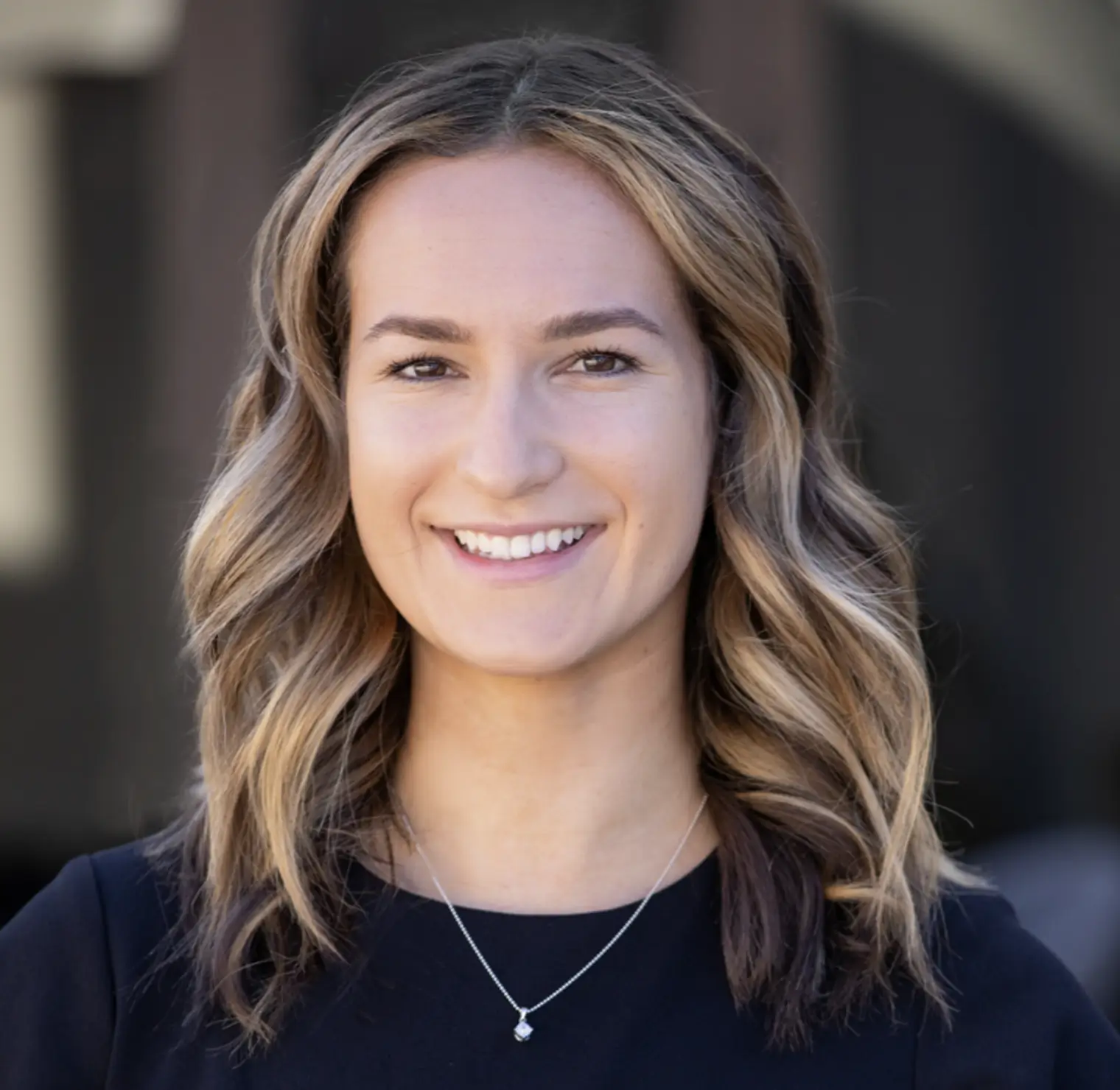 Smiling woman with wavy blonde hair wearing a black top and a silver necklace with a pendant.