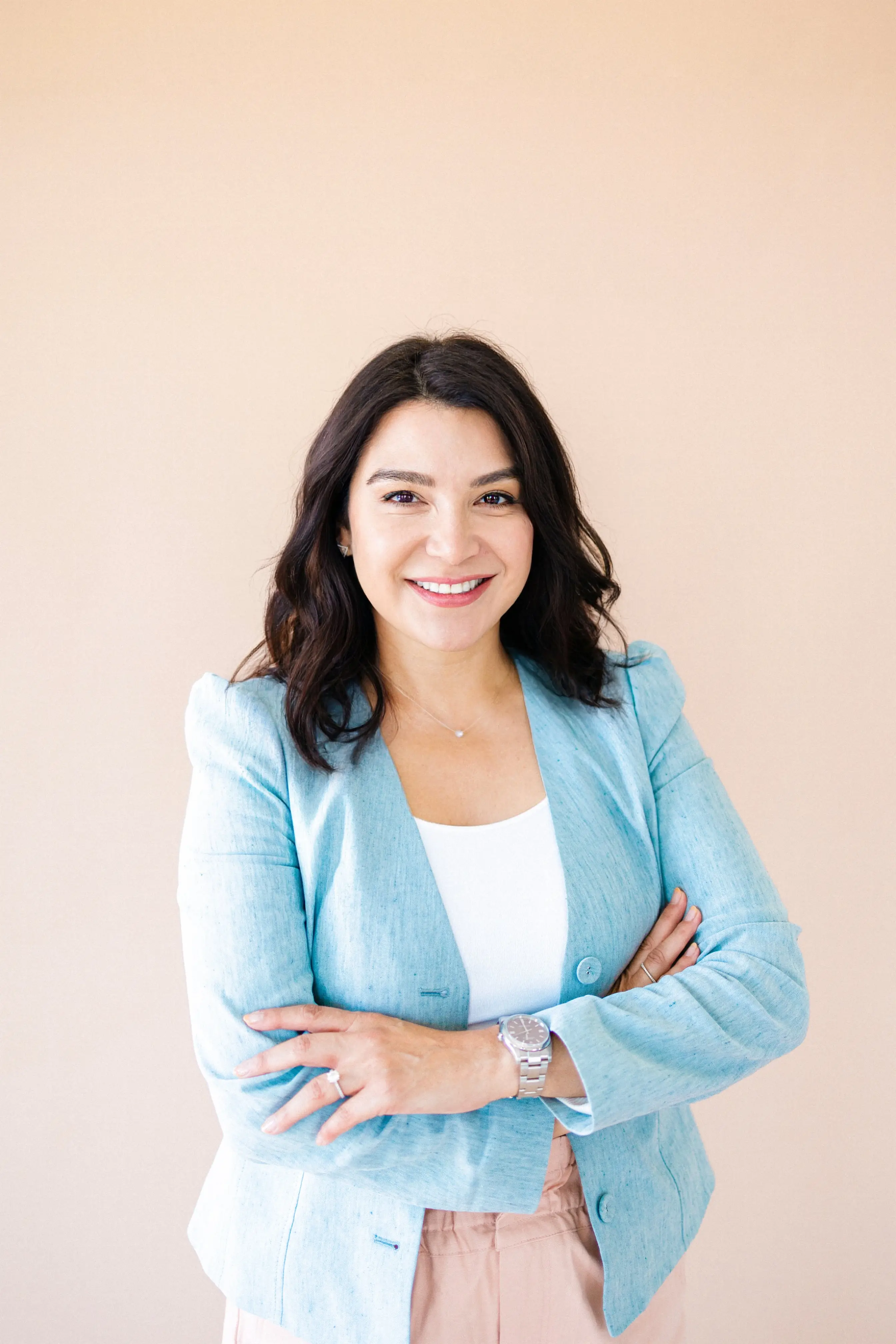 Smiling woman with dark hair wearing a light blue blazer, white top, and beige pants with arms crossed against a beige background.