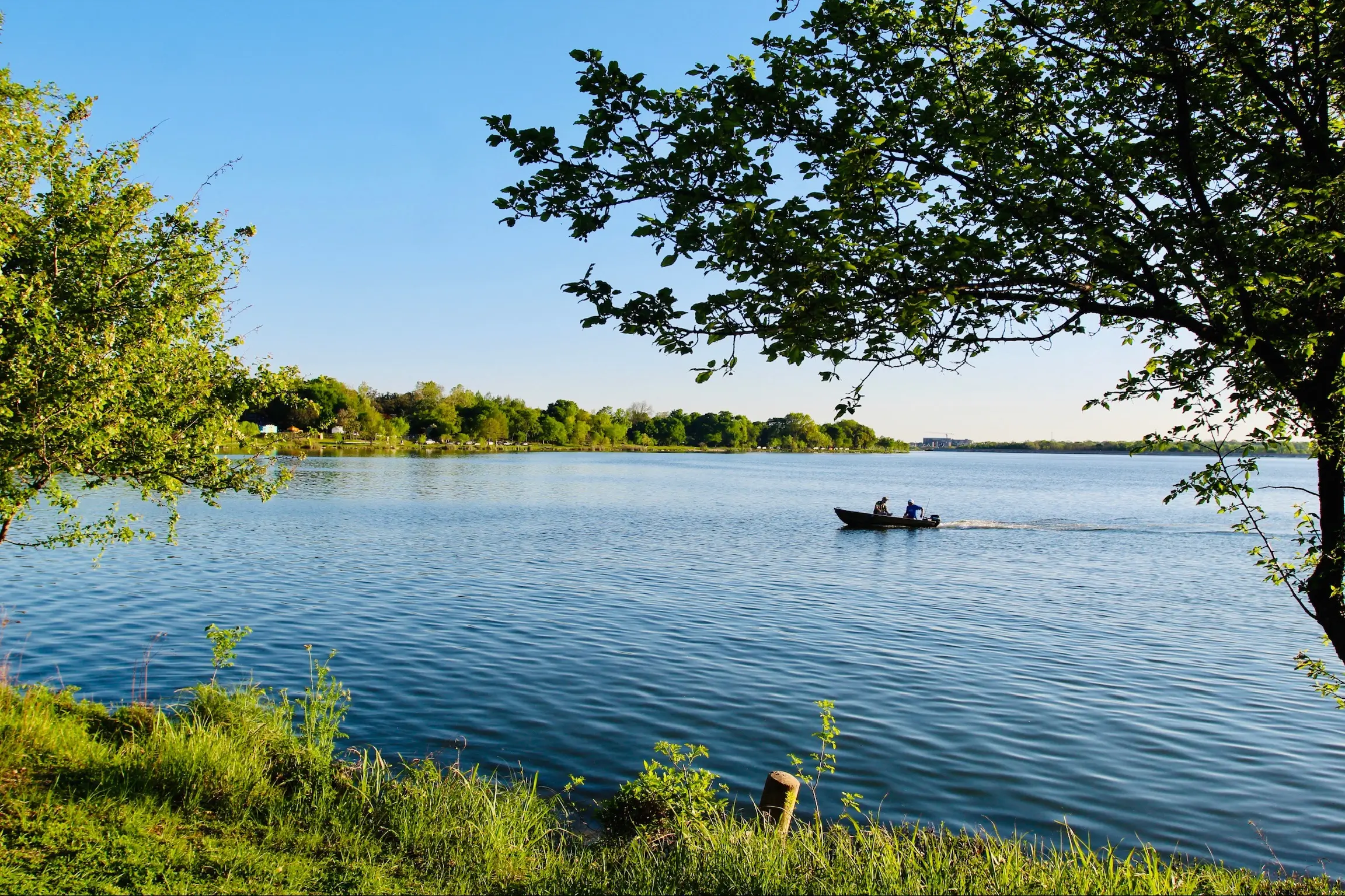 Two people on a small motorboat on a calm lake surrounded by green trees under a clear blue sky.