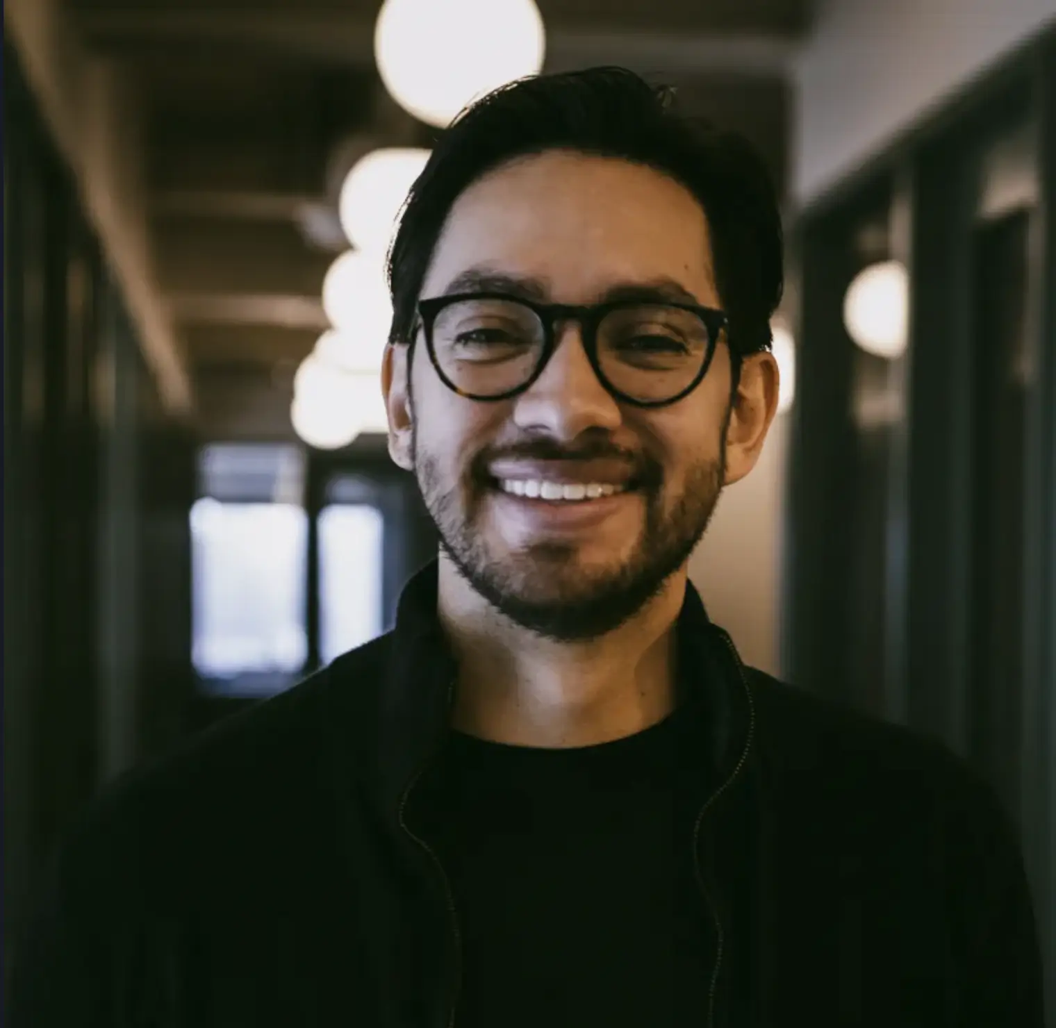 Smiling man with glasses and beard standing in a dimly lit hallway with round ceiling lights.