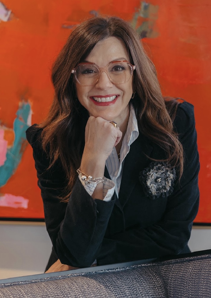 Smiling woman with long brown hair, wearing glasses and a dark blazer, posing with her hand under her chin against a vibrant red abstract background.