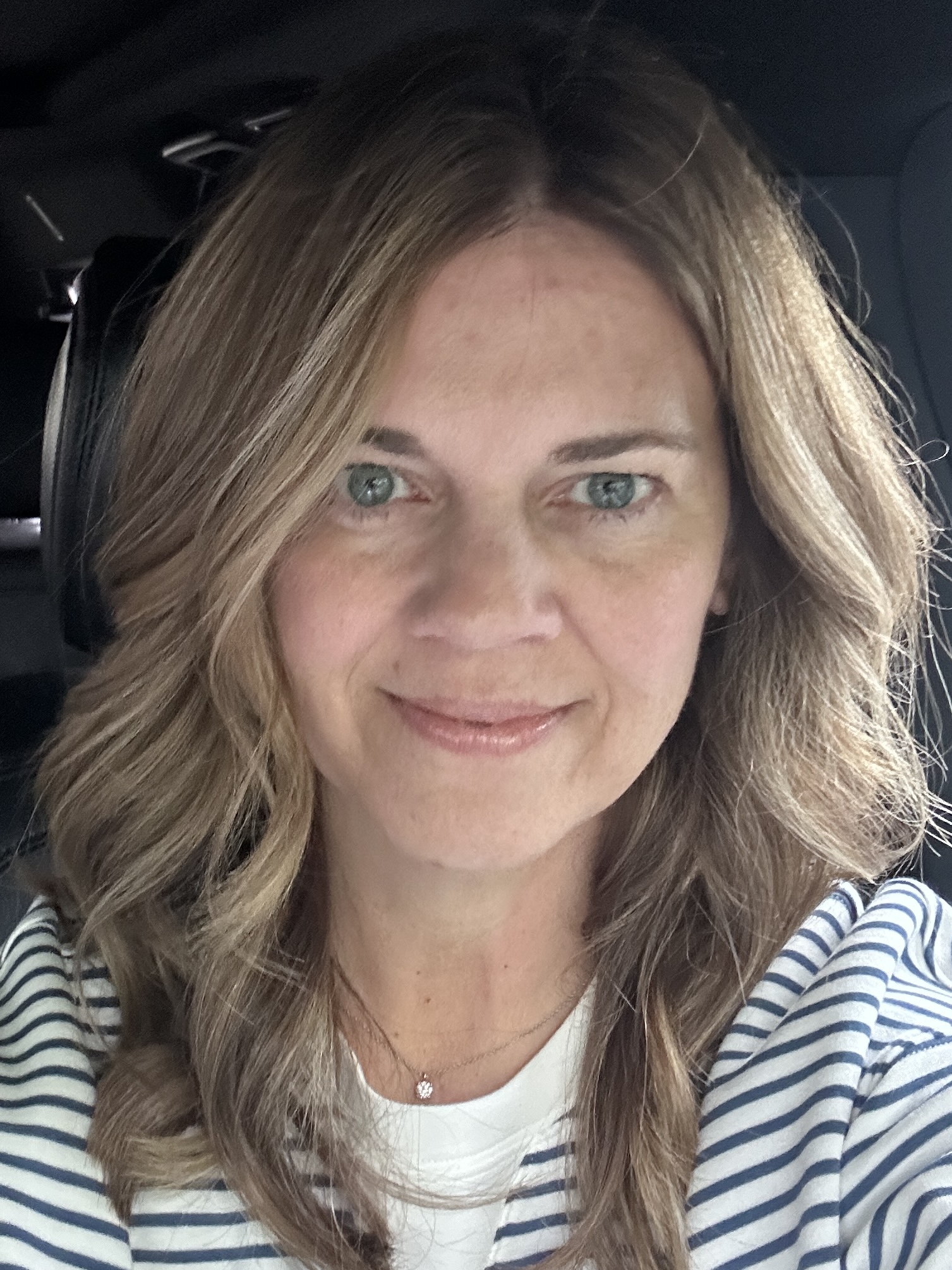 Woman with wavy light brown hair wearing a striped shirt and a small necklace, smiling inside a car.