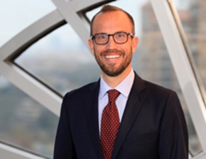 Smiling man with glasses wearing a dark suit and red tie standing indoors by a window with a blurred cityscape background.