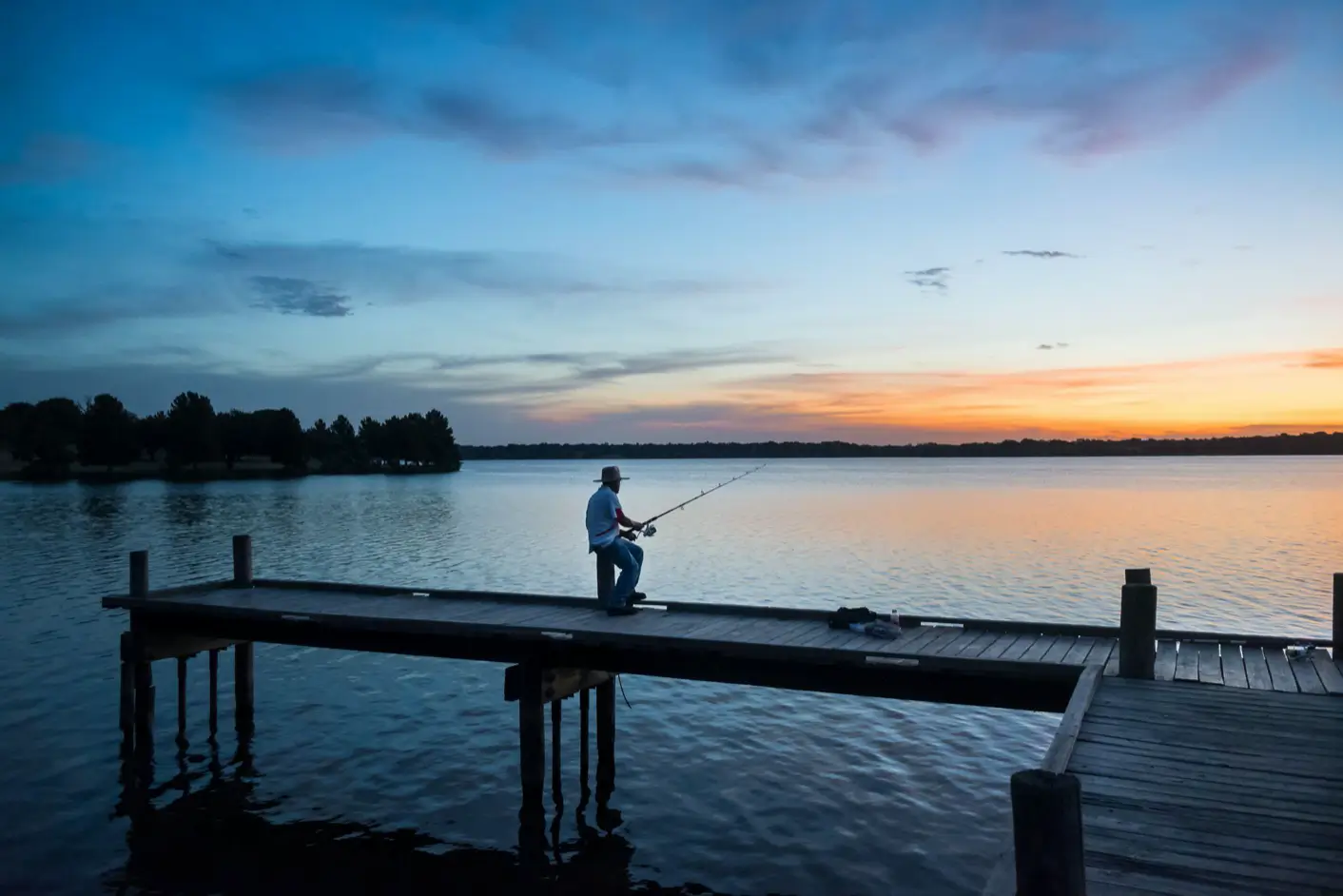 Person fishing on a wooden dock at sunset with a calm lake and distant tree-lined shore on White Rock Lake.